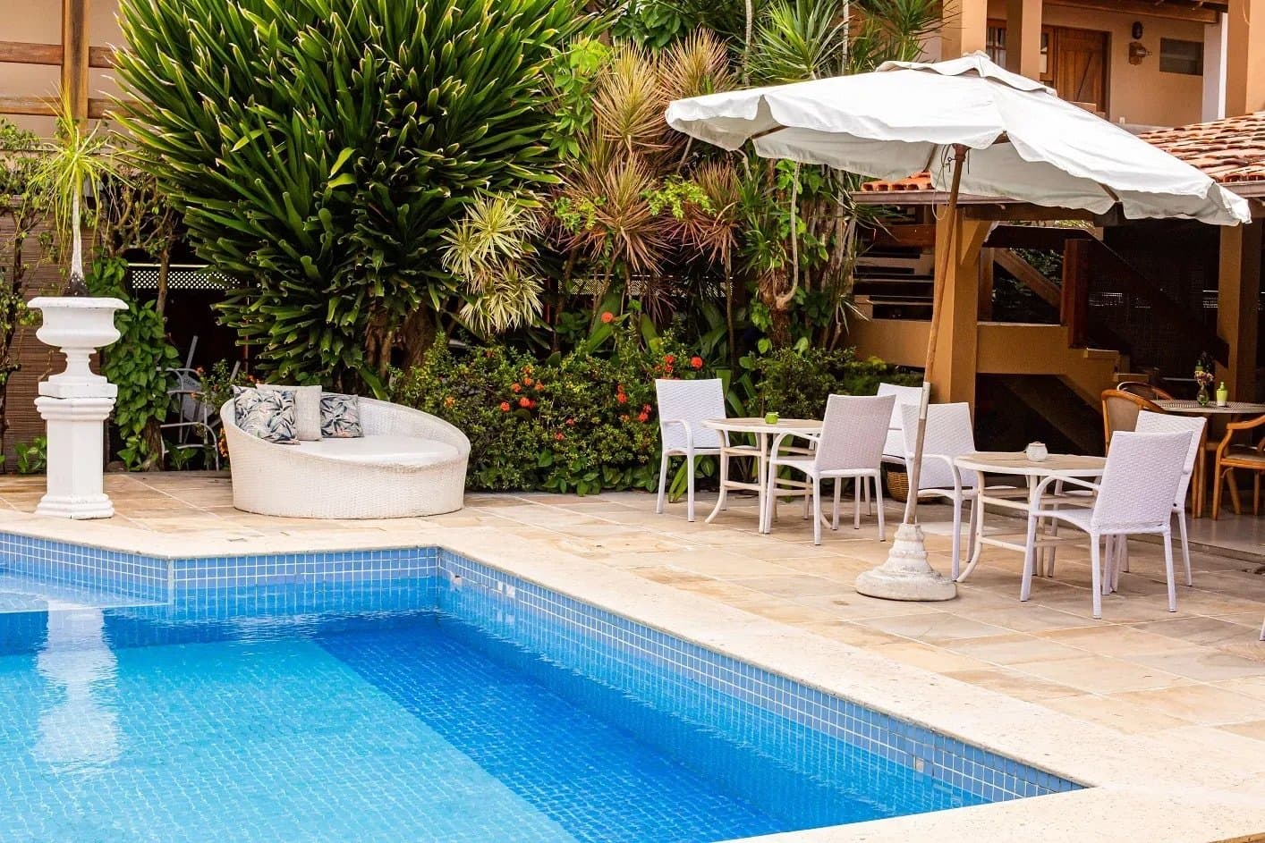 Pousada com piscina no fim do dia: recompensa justa, sem discussão. Área de piscina ensolarada em uma pousada na rota de ciclismo na Bahia, com sofá de vime branco, mesas ao ar livre sob guarda-sol e jardim tropical exuberante