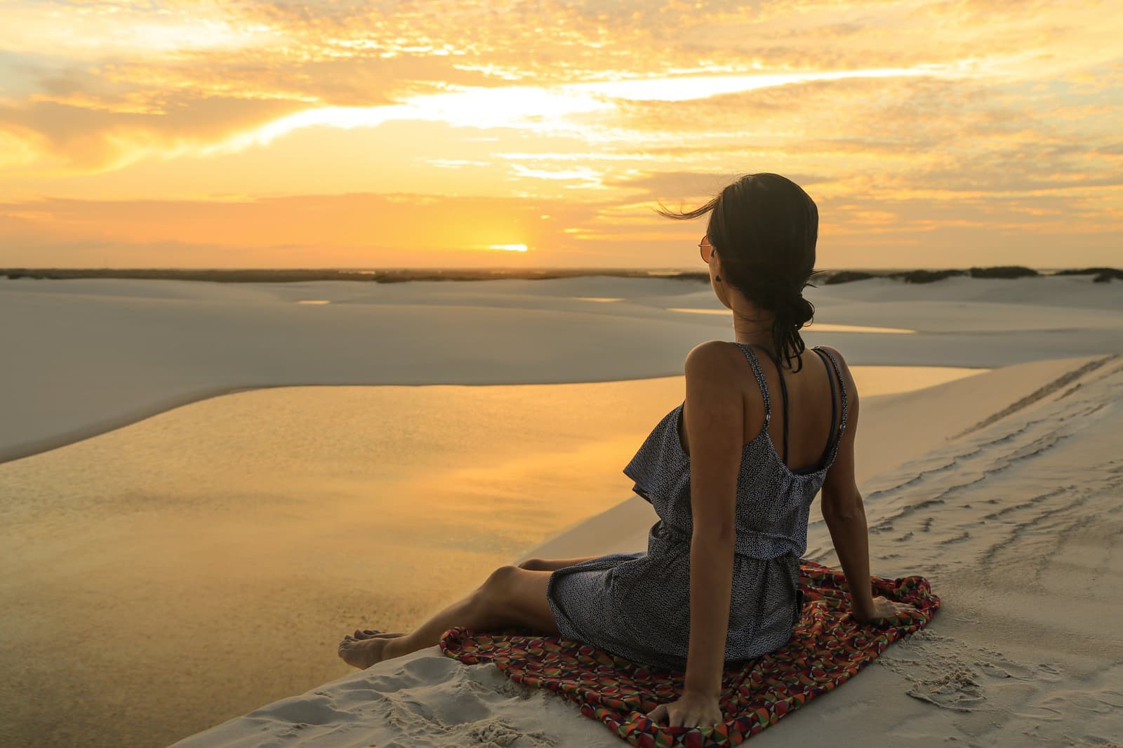 Tem hora que a gente para, senta na duna e deixa o sol fazer o resto. Uma mulher sentada em uma manta colorida no topo de uma duna de areia branca contempla um pôr do sol dourado sobre a paisagem dos Lençóis Maranhenses