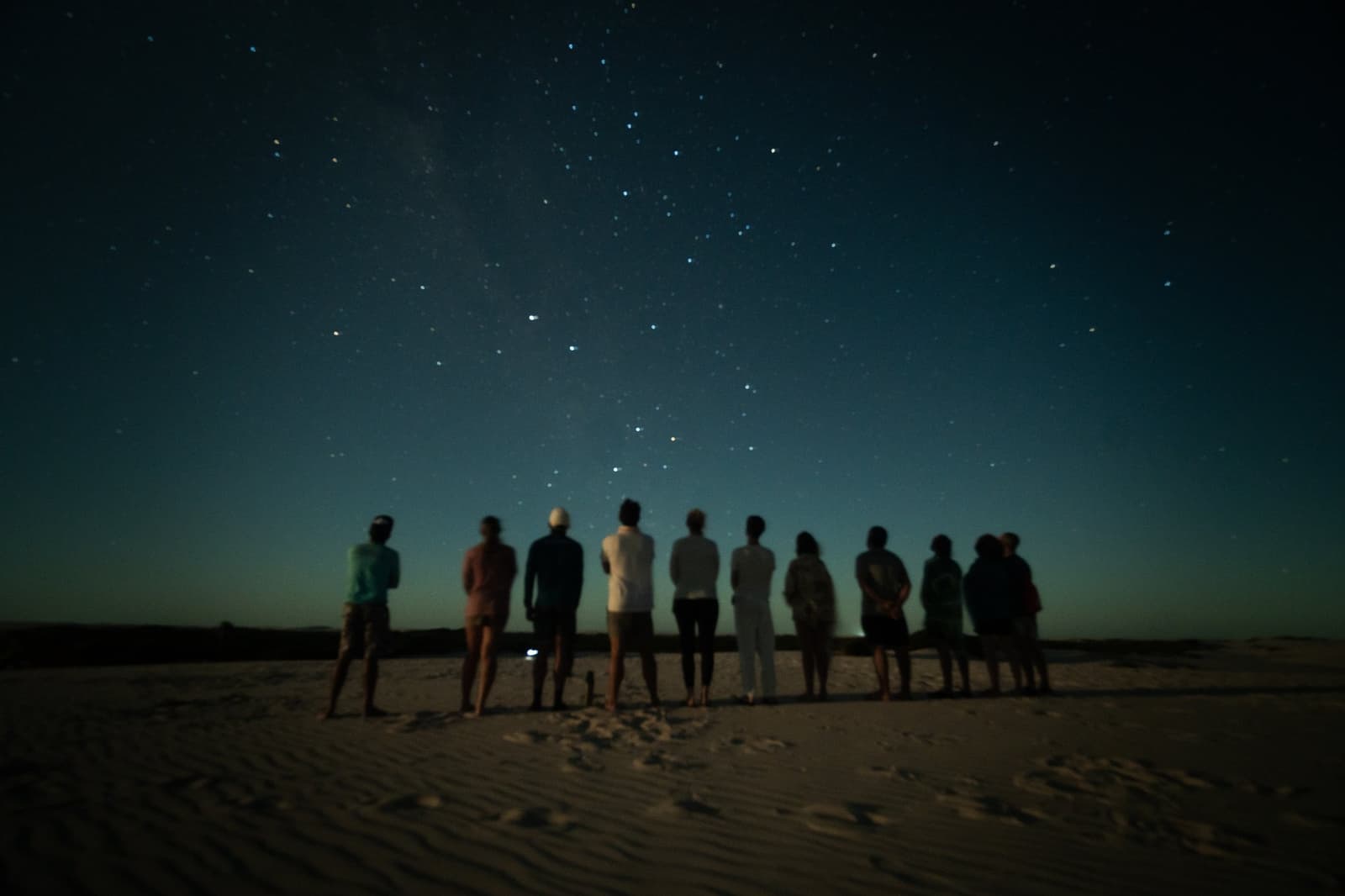 Dez pessoas, zero luz artificial, e o universo inteiro aparecendo para a festa. Um grupo de viajantes enfileirado sobre as dunas brancas durante o trek pelos Lençóis Maranhenses saindo de Atins, em silhueta contra o céu noturno azul profundo repleto de estrelas e um tênue traço da Via Láctea