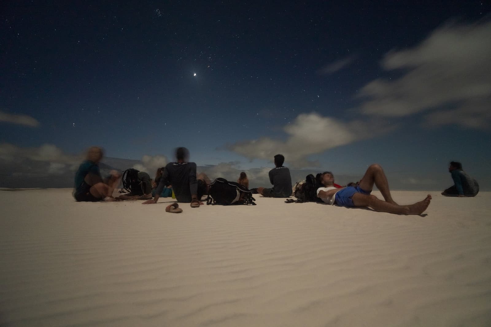 A melhor sala de estar do Brasil não tem paredes. Um grupo de viajantes deitados e sentados sobre dunas de areia branca sob um céu noturno azul-escuro durante o trek pelos Lençóis Maranhenses saindo de Atins, com estrelas e nuvens esparsas visíveis acima