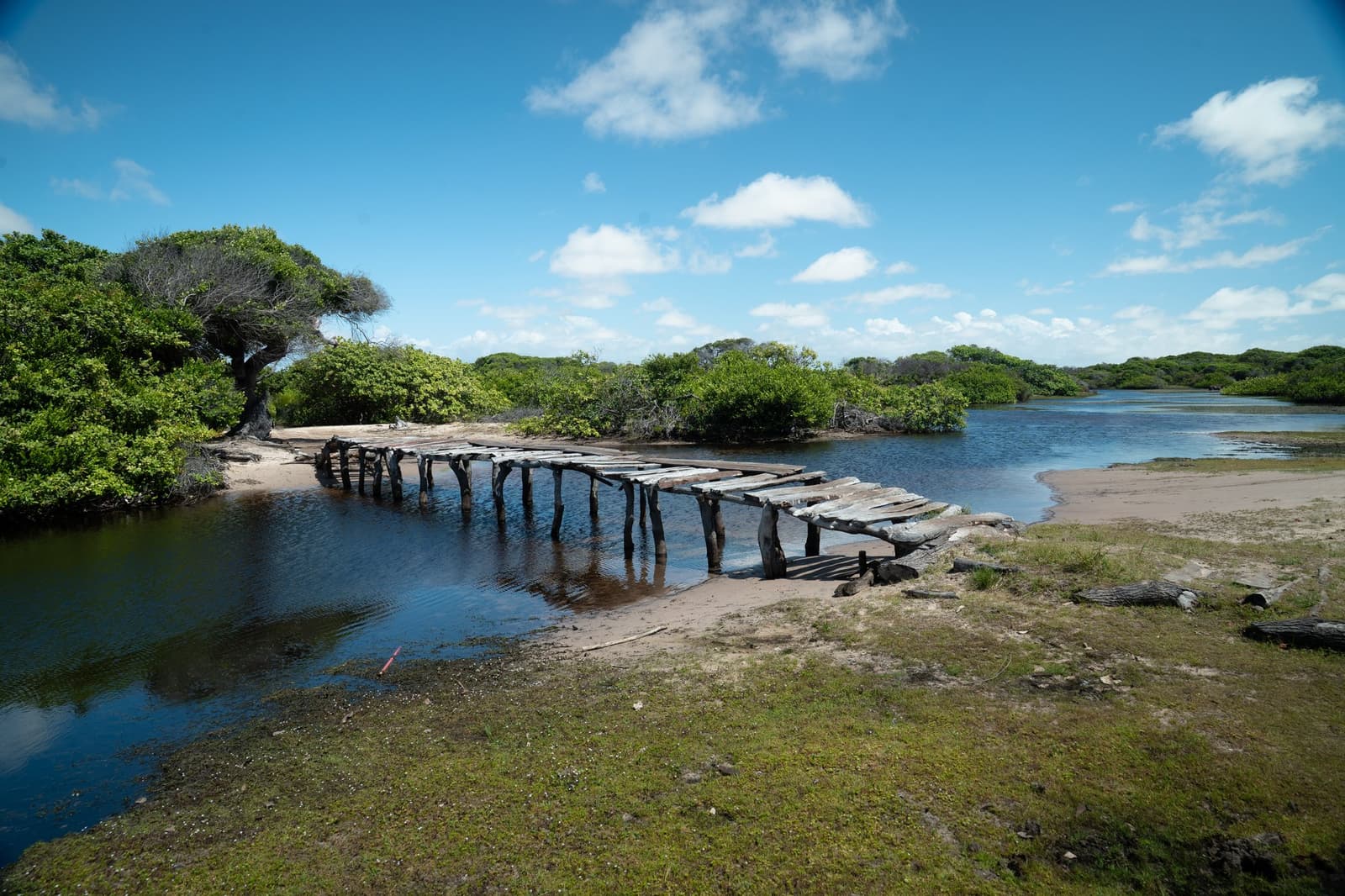 Tábua por tábua, alguém insistiu em conectar as duas margens. Uma passarela de madeira desgastada atravessa um canal raso ladeado por vegetação costeira baixa e margens arenosas no percurso do trek pelos Lençóis Maranhenses, sob um céu azul com nuvens esparsas.