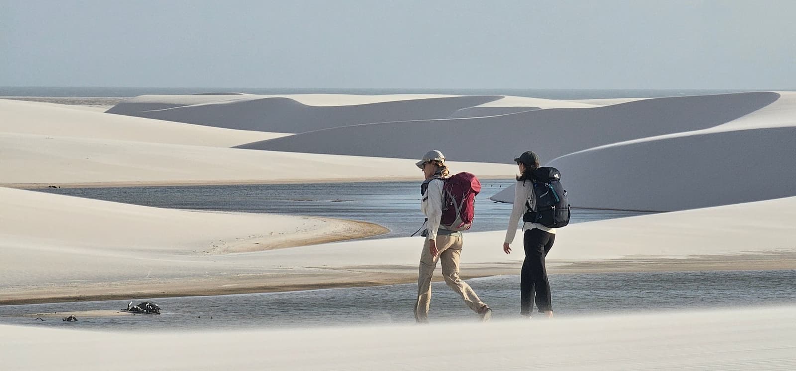 Uma boa prosa e as dunas parecem passar mais rápido. Dois caminhantes com mochilas atravessando as dunas brancas do trek pelos Lençóis Maranhenses, com uma lagoa rasa e longas cristas de dunas se estendendo até o horizonte ao fundo
