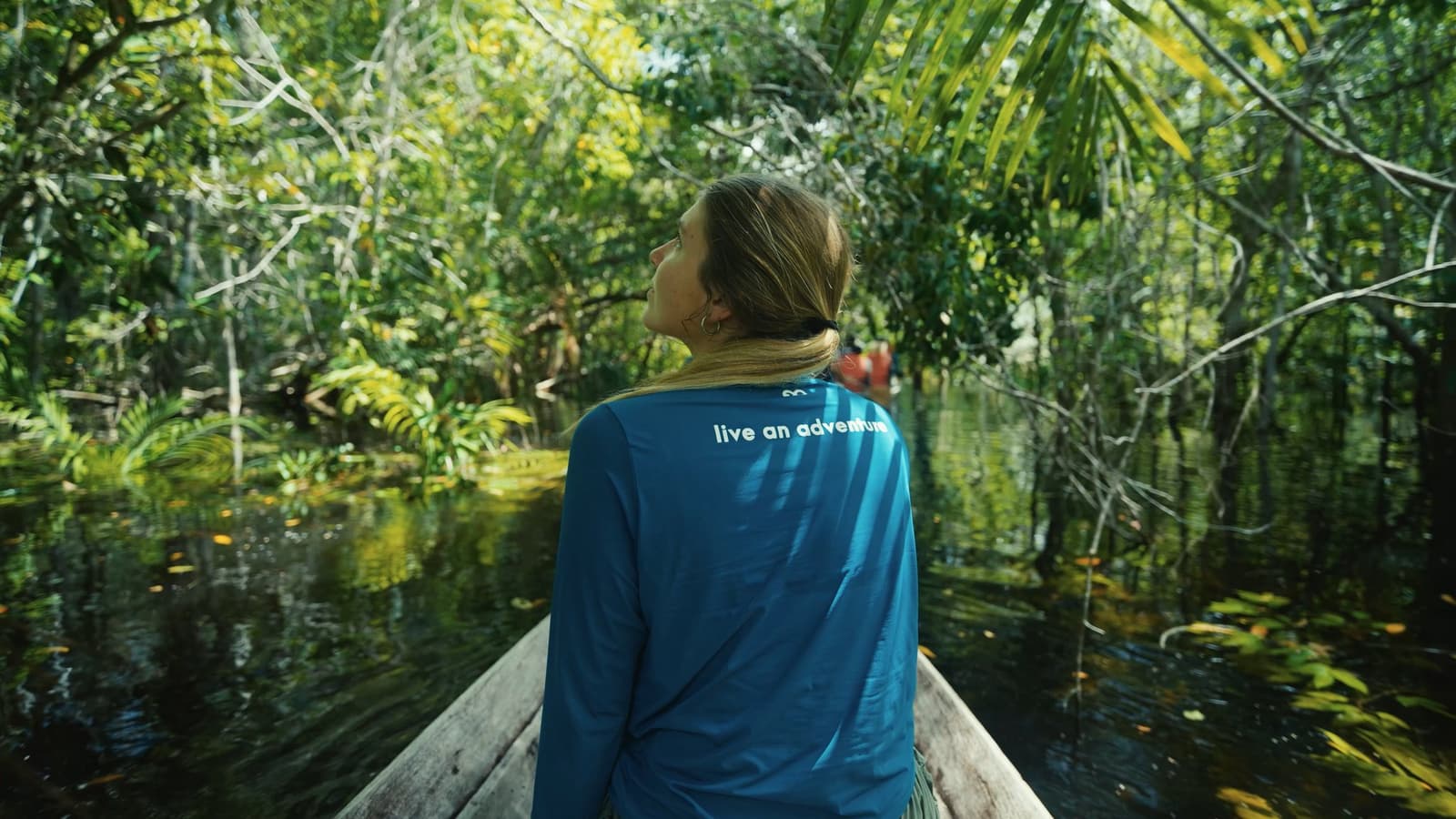 On n'explore pas l'Amazonie, on se laisse absorber par elle. Une voyageuse assise à la proue d'une pirogue en bois lève les yeux vers la forêt inondée d'igapó lors d'une excursion au Mirante do Gavião Amazon Lodge, l'eau sombre aux tanins reflétant la canopée verdoyante