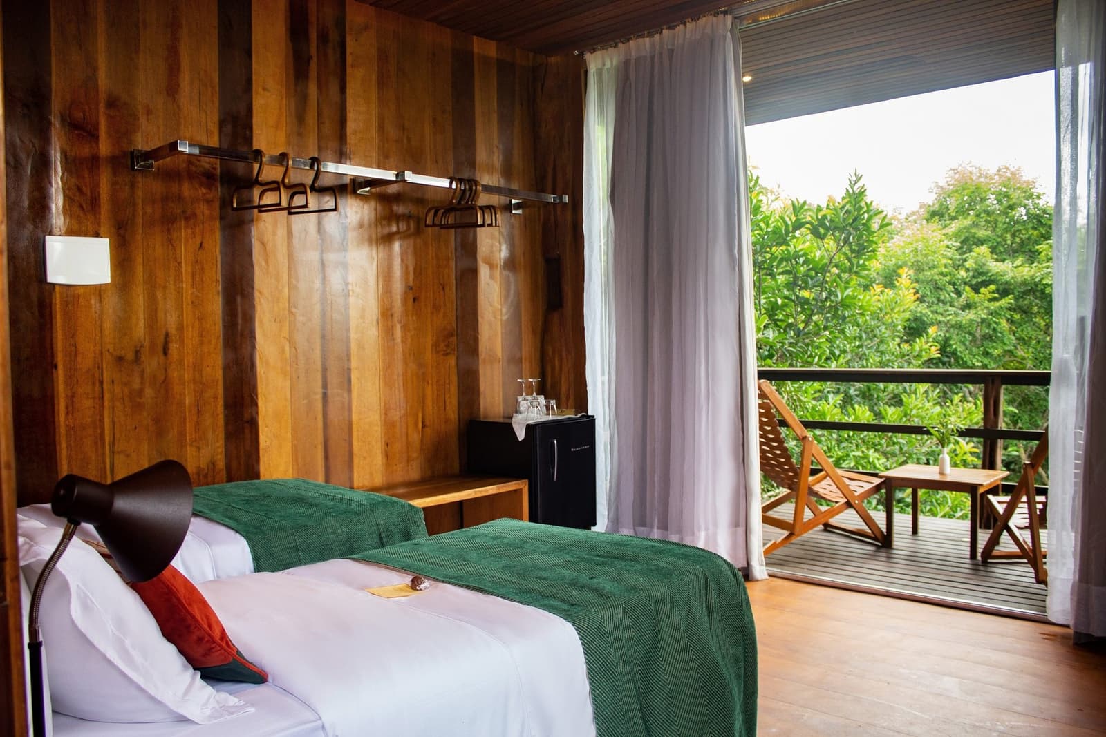 Le rideau tiré, c'est la forêt. Le rideau ouvert, c'est encore la forêt. Chambre lambrissée de bois au Mirante do Gavião avec un lit bien fait, un mini-réfrigérateur et des portes vitrées ouvertes sur un balcon privé surplombant une dense canopée amazonienne