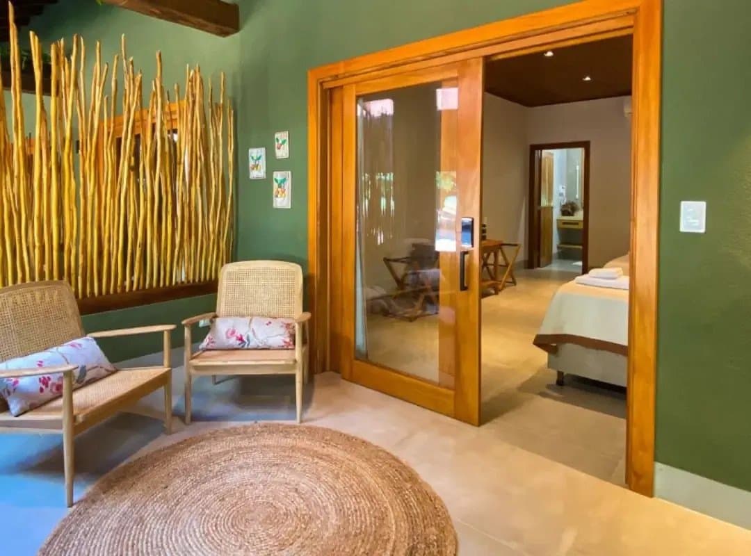 Des matières naturelles, une palette verte, et ce silence rare qu'on savoure. Coin salon d'un lodge de safari dans le sud du Pantanal, avec des fauteuils en rotin, un tapis rond tressé, des gravures botaniques, un mur décoratif de branches dorées séchées et une porte coulissante en verre ouvrant sur une chambre avec un lit bien fait