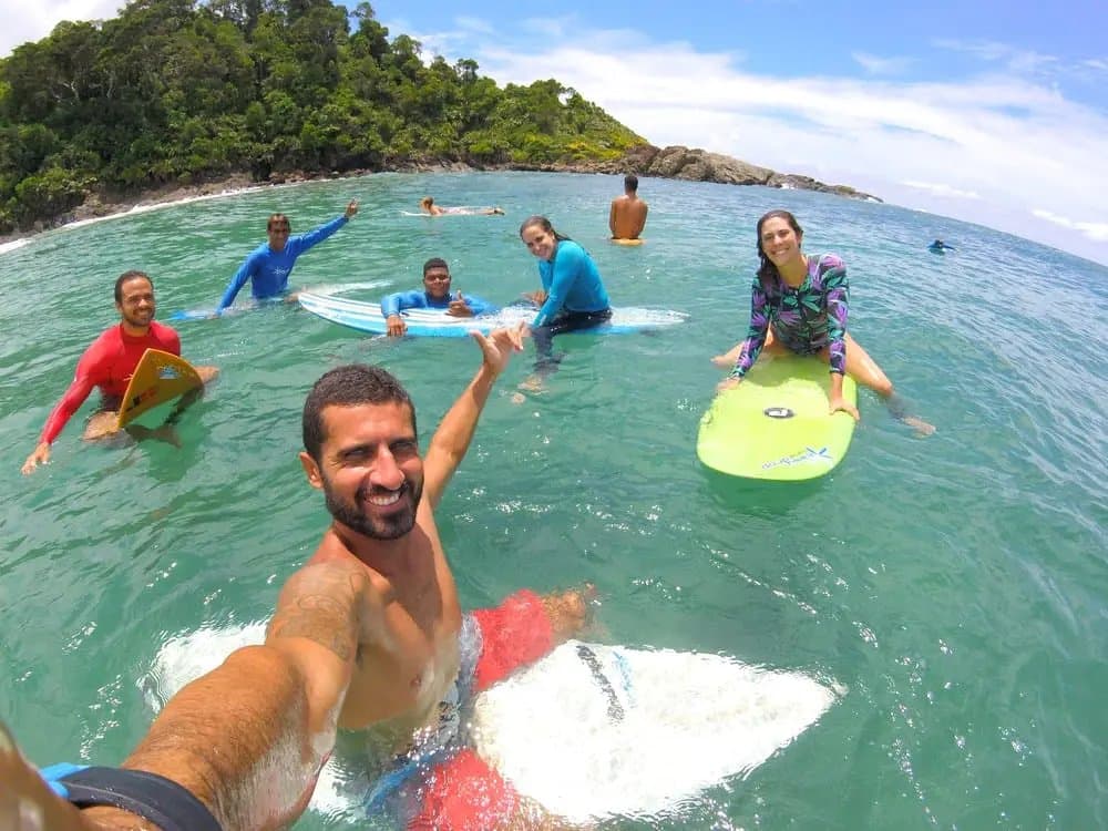 Vijf boards, volop lachen, en niemand die aan morgen denkt. Een lachende instructeur maakt een groothoek water-selfie met een groep studenten tijdens surflessen in Itacaré, allemaal met kleurrijke surfboards in helder turquoise water, met een beboste kaap op de achtergrond.