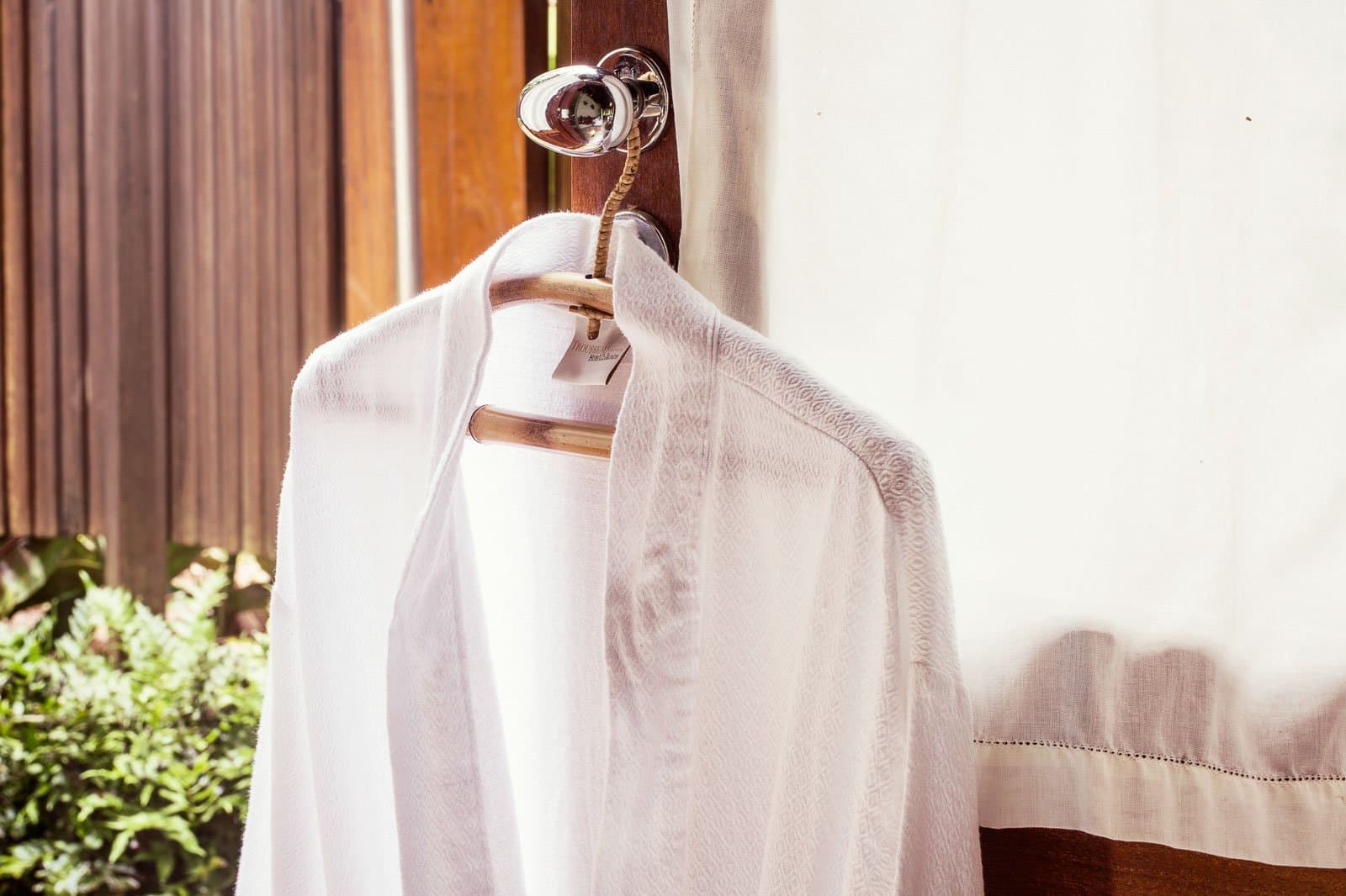 Le peignoir prêt, la forêt qui attend juste derrière. Photo: | SamuelMelim Photography Un peignoir en éponge blanche et un vêtement blanc texturé suspendus à un cintre en bois accroché à une porte sombre, avec une végétation tropicale visible par la fenêtre dans une chambre de lodge pour l'observation des oiseaux en forêt amazonienne