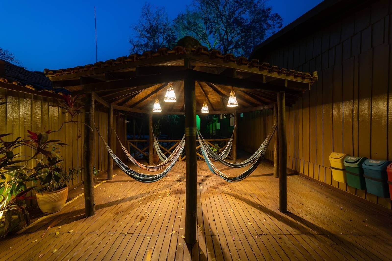 Der Platz, an dem der Wecker von morgen plötzlich ganz weit weg klingt. Überdachte Holzterrasse in der Dämmerung auf einer Budget-Pantanal-Tour, mit mehreren bunten Hängematten unter warmen Pendelleuchten und einem tiefblauen Abendhimmel über den Baumkronen