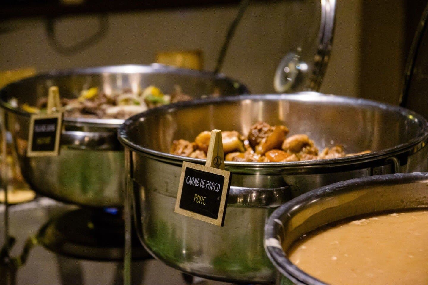 Kräftig, warm, unkompliziert, genau das Richtige nach einem langen Tag draußen. Warmhaltebehälter aus Edelstahl an einem Lodge-Büfett, der vordere Topf mit der Aufschrift 'Carne de Porco' gefüllt mit geschmortem Schweinefleisch, Teil der Vollpension bei dieser Pantanal-Reise für kleines Geld