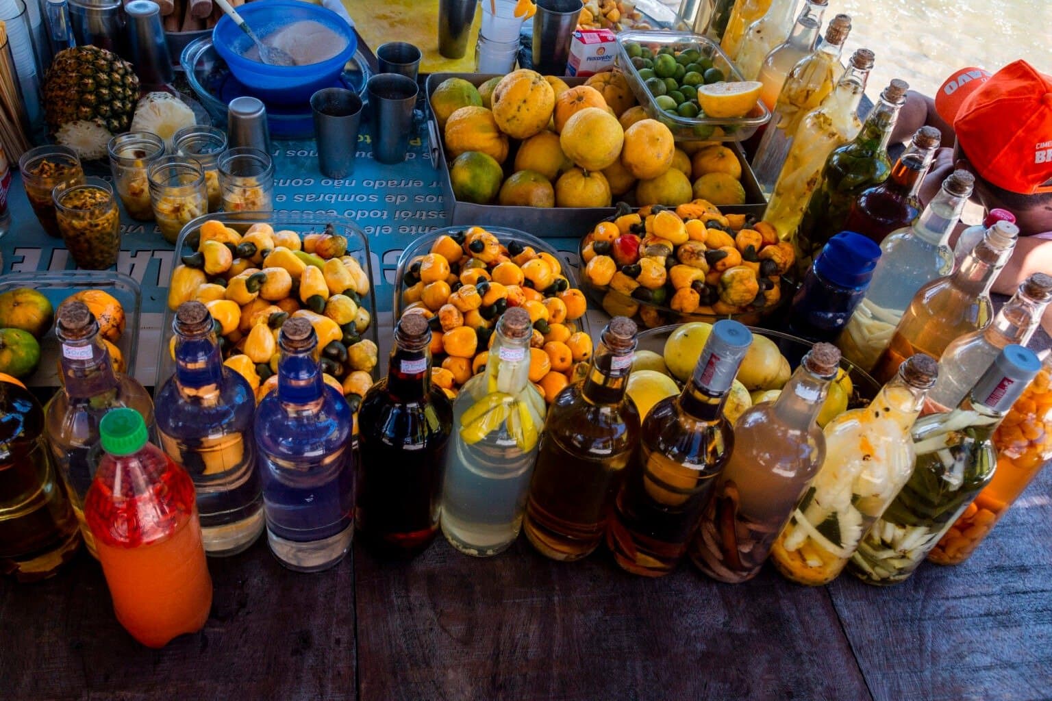 Escolha uma garrafa, qualquer uma. Essas receitas têm história e capricho por trás. Uma banca de mercado repleta de bandejas de caju fresco, frutas cítricas tropicais e fileiras de garrafinhas coloridas de licor artesanal, parte da cultura gastronômica local no roteiro completo de Lençóis Maranhenses