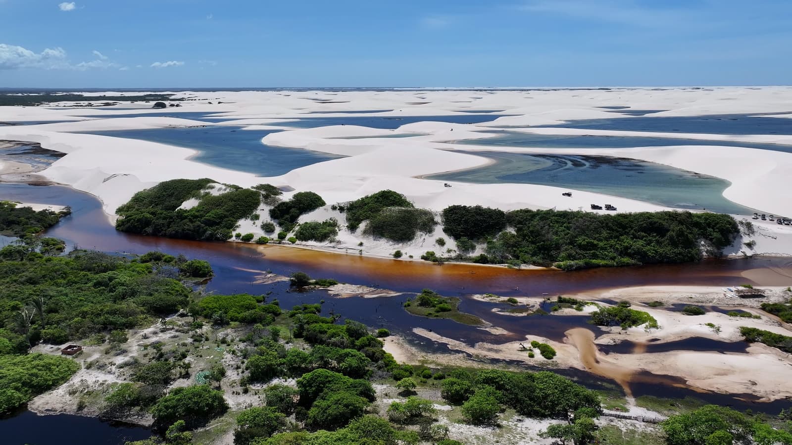Rio preto, areia branca, lagoa azul, os Lençóis jogam com as cores sem pedir licença. Vista aérea de um rio de águas escuras e tânicas serpenteando pela vegetação verde antes de encontrar as dunas brancas e lagoas turquesa do roteiro completo pelos Lençóis Maranhenses