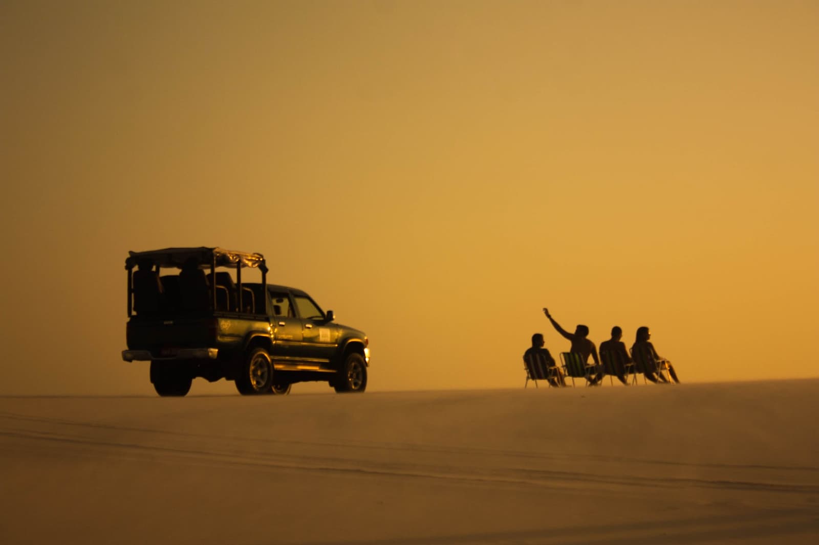 Cadeiras nas dunas, o pôr do sol faz o resto. Quatro viajantes sentados em cadeiras de praia sobre uma ampla duna ao entardecer dourado, ao lado de um veículo 4x4 em silhueta durante o roteiro completo pelos Lençóis Maranhenses