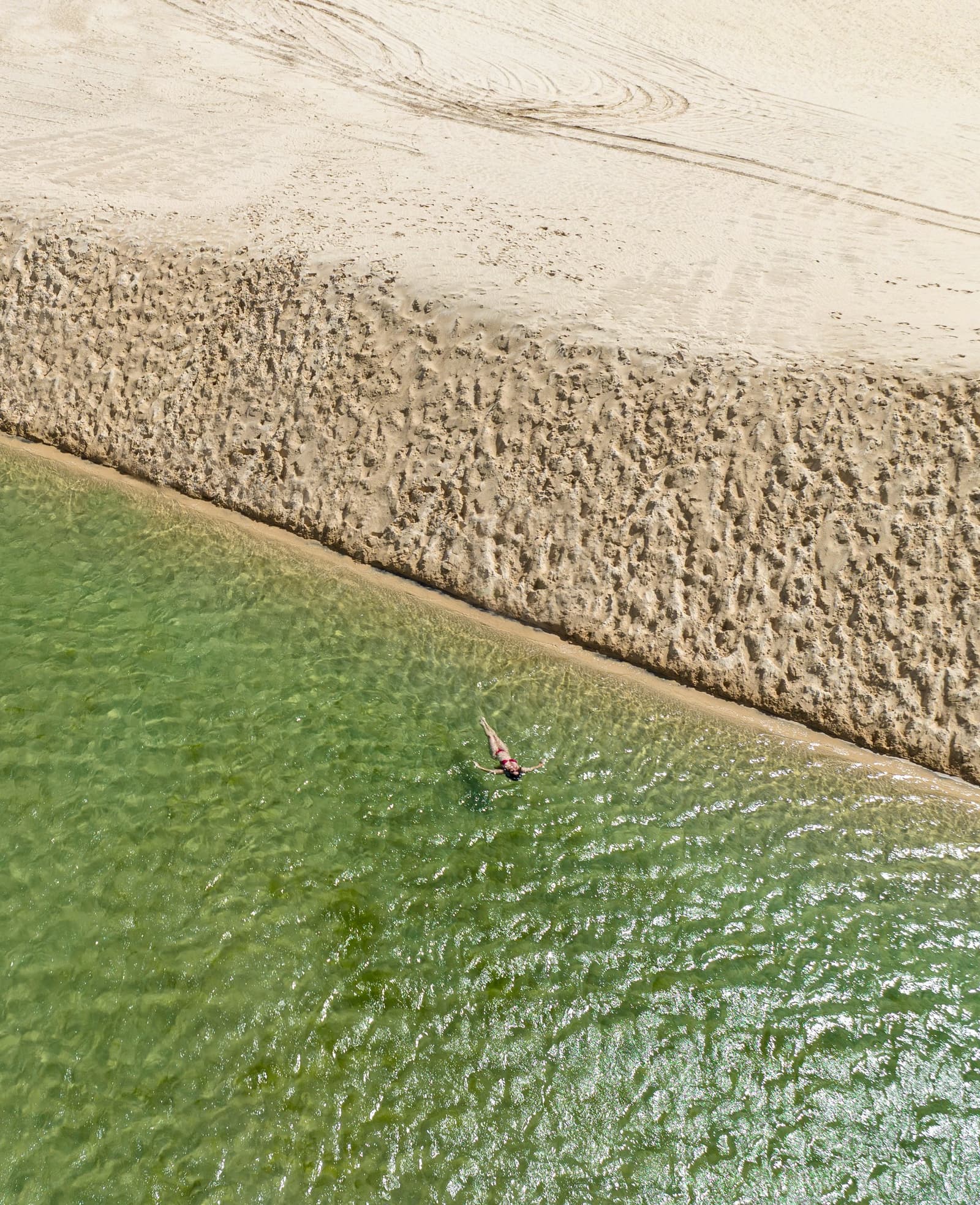 Água verde, areia branca e nenhuma pressa, os Lençóis têm esse efeito. Vista aérea de uma pessoa sozinha flutuando em água verde e cristalina ao lado de um banco de areia texturizado no trek pelos Lençóis Maranhenses saindo de Atins