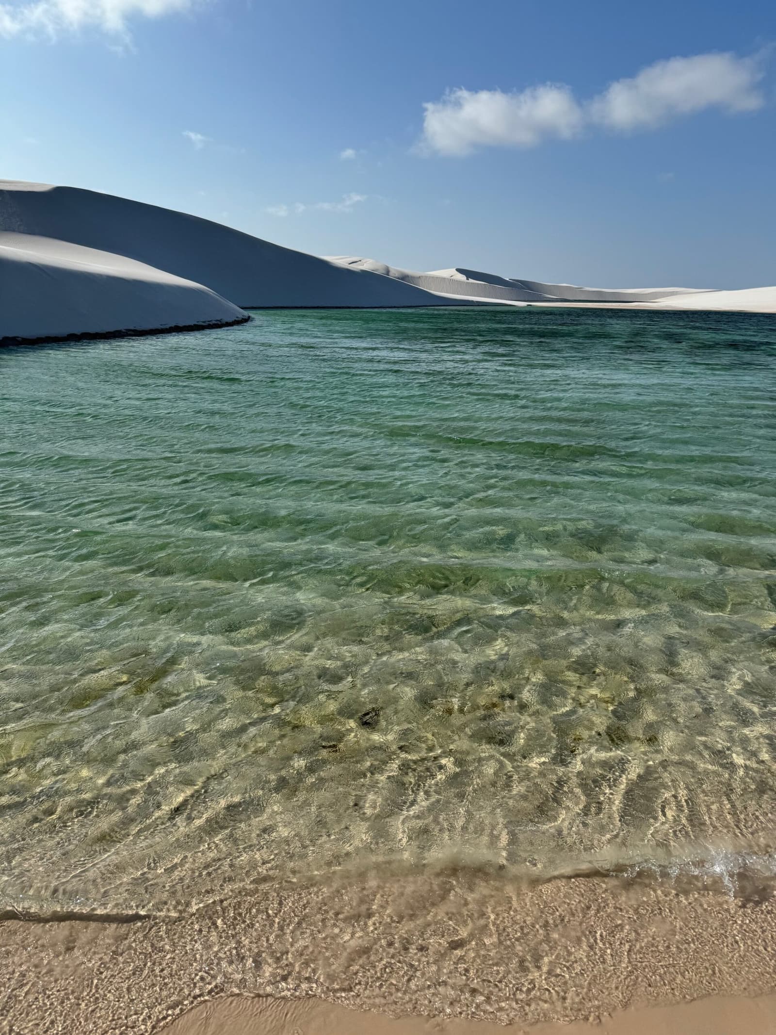 Água gelada, areia branca e nenhuma fila para entrar. Água verde e cristalina de uma lagoa de água doce no trek pelos Lençóis Maranhenses, com altas dunas de areia branca na margem oposta sob um céu azul