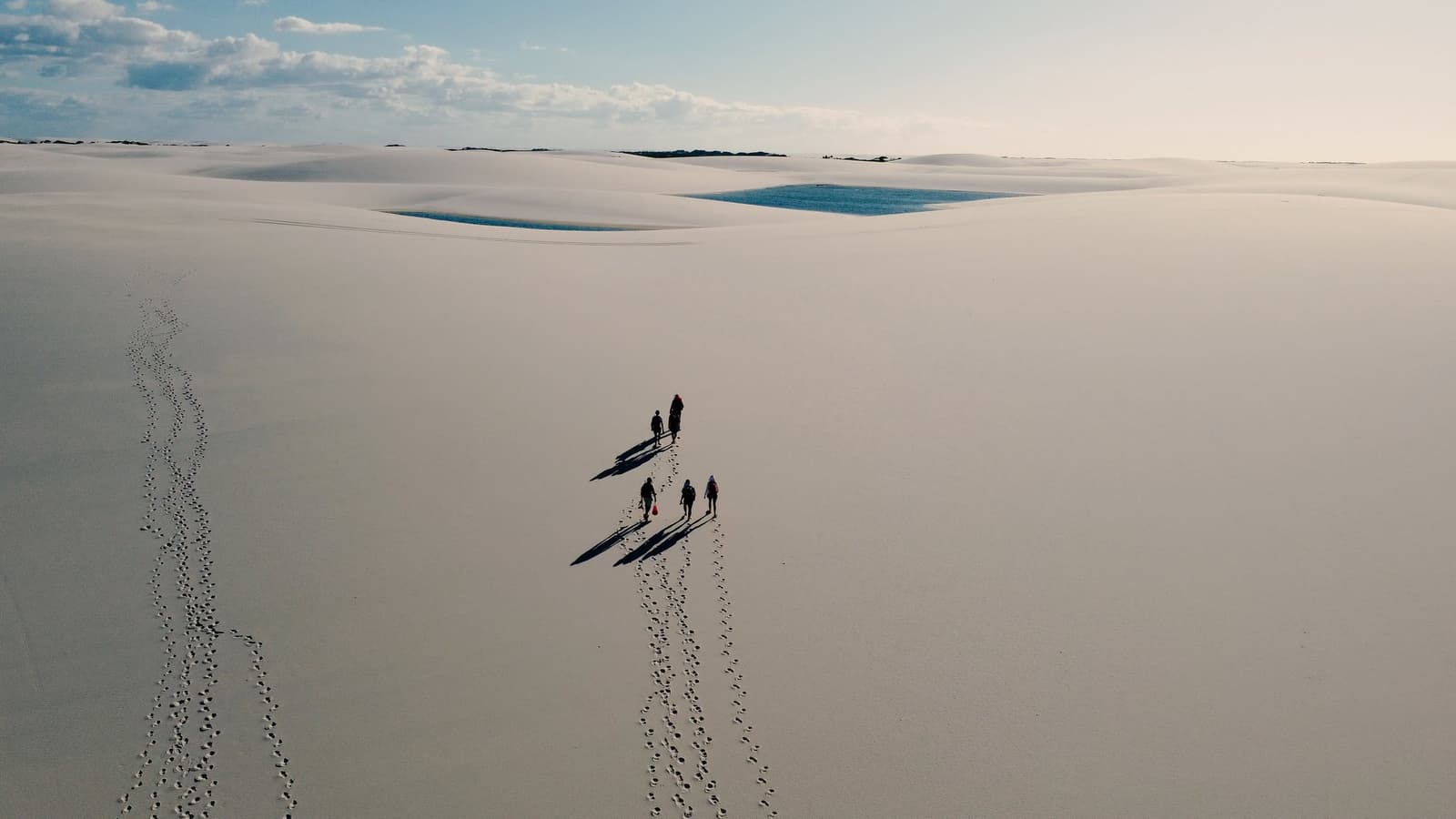 Quatre silhouettes dans l'immensité blanche, et des traces que le vent effacera bientôt. Vue aérienne de quatre randonneurs marchant sur de vastes dunes de sable blanc lors d'un trekking dans les Lençóis Maranhenses, avec des lagunes bleues visibles au loin et de longues ombres derrière eux