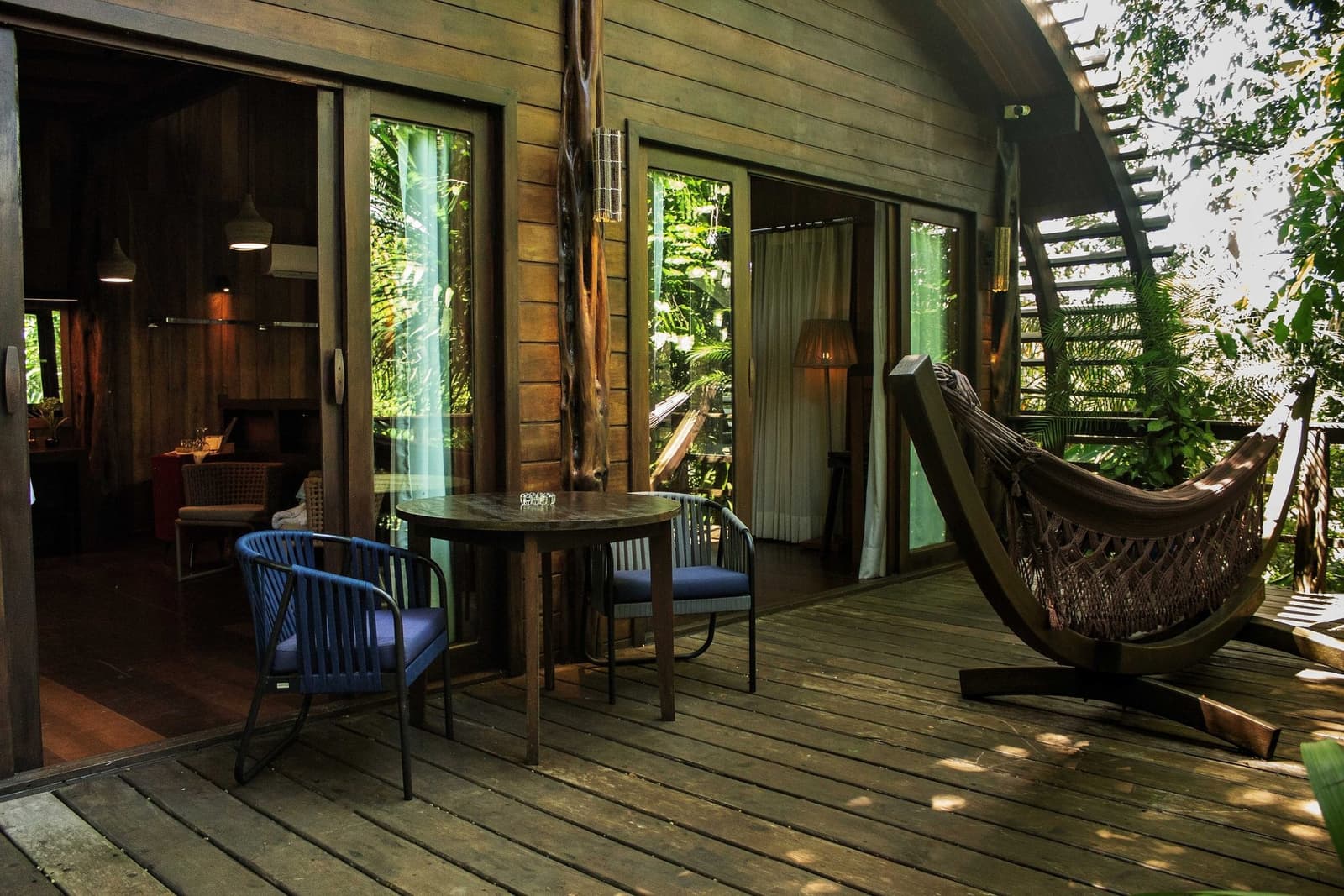 Le hamac ou la table. Un dilemme qu'on accepte volontiers. Une terrasse en bois au Mirante do Gavião avec une table ronde, deux fauteuils rembourrés, un hamac tressé sur un support courbé et des portes coulissantes en verre ouvrant sur une chambre chaleureusement éclairée entourée d'une végétation dense