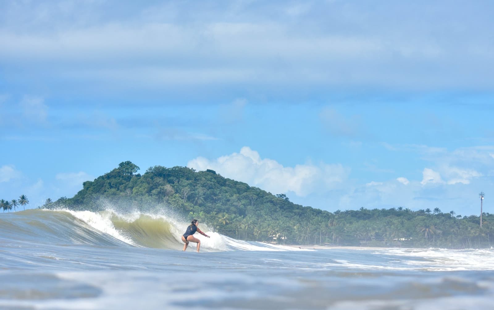 Een mooie golf, niemand in de weg. Dit is precies waarvoor je komt. Photo: Picasa Een vrouw hurkt laag op haar surfboard tijdens surfles in Itacaré, terwijl een perfect brekende golf rolt voor een weelderig beboste kaap onder een blauwe hemel.