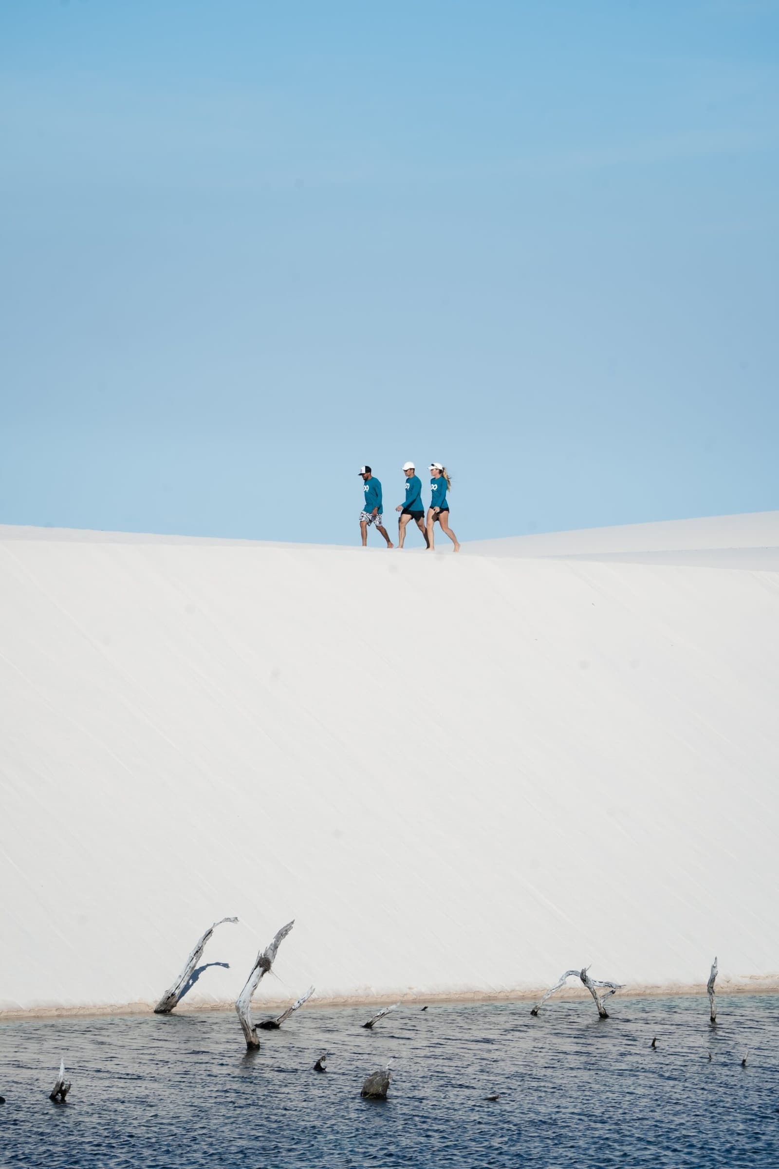 O branco da areia, o azul da lagoa, e você lá no meio disso tudo. Três trekkers com camisas azul-petróleo caminham pelo cume de uma duna de areia branca no trek dos Lençóis Maranhenses saindo de Atins, com uma lagoa escura ladeada por galhos submersos em primeiro plano e céu azul limpo ao fundo
