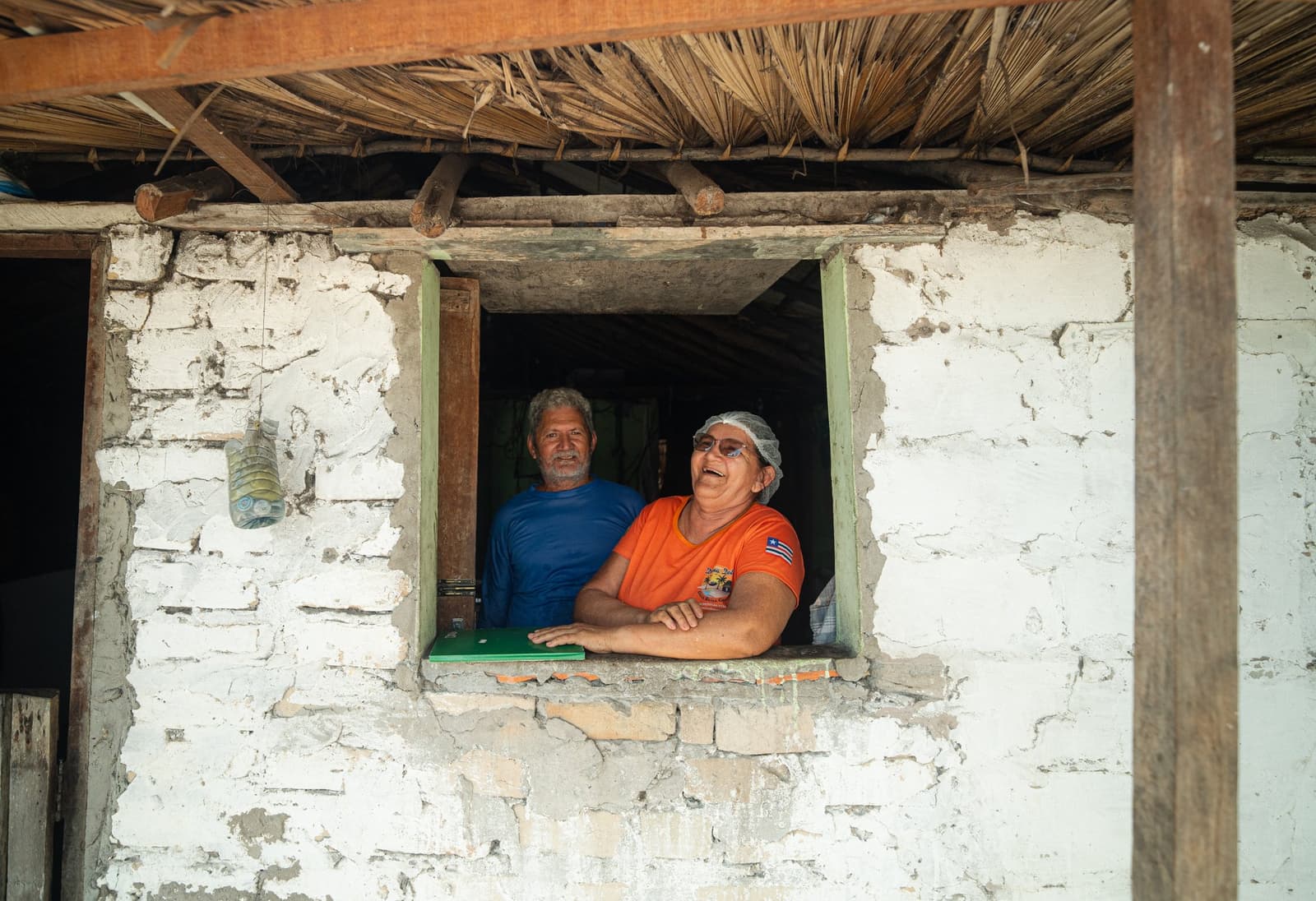 Os vizinhos sempre sabem das novidades antes de todo mundo. Um homem de camiseta azul e uma mulher de camiseta laranja com touca de cabelo se debruçam sorridentes numa janela de casa de tijolos caiados com telhado de palha, representando a hospitalidade do trek Lençóis Maranhenses.