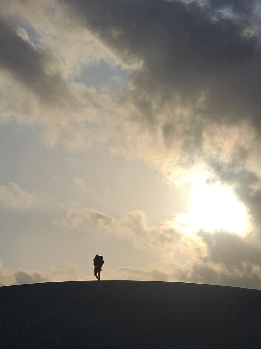 Uma figura, uma duna, um céu enorme. O resto fica para depois. Silhueta de um caminhante com mochila no topo de uma duna de areia branca durante o trek pelos Lençóis Maranhenses, sob um céu nublado dramático com o sol rompendo as nuvens
