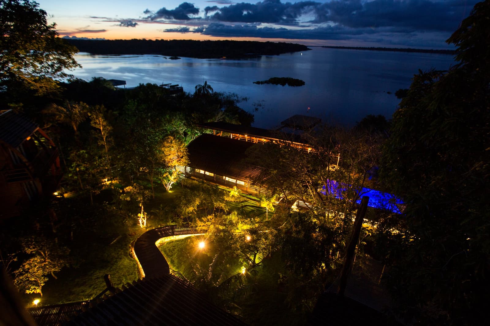 L'heure bleue sur le fleuve, et le lodge qui s'allume doucement dans la canopée. Vue aérienne au crépuscule du Mirante do Gavião avec un éclairage doré dans les jardins, une promenade en bois sinueuse, une piscine illuminée en bleu et le vaste Rio Negro s'étendant jusqu'à l'horizon