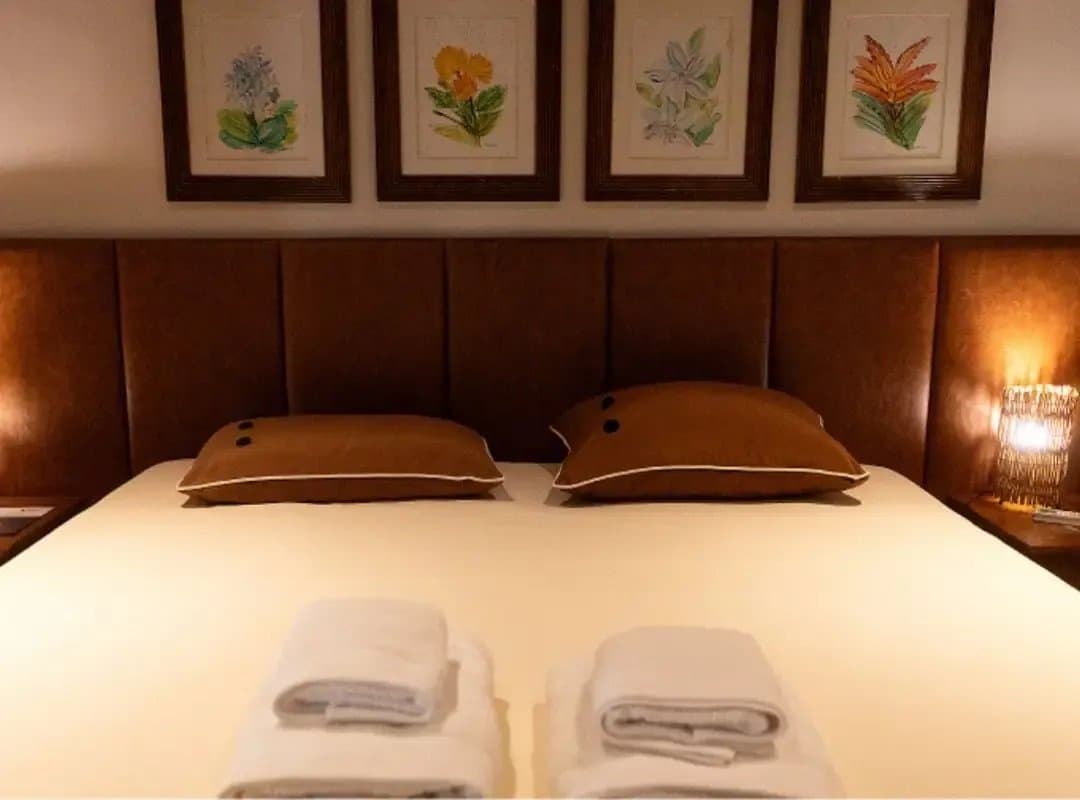 Gravures botaniques, lumière douce, serviettes déjà pliées. Le repos fait partie du voyage. Lit double bien fait avec des coussins couleur terre cuite, des serviettes blanches pliées au pied du lit, une lampe de chevet chaleureuse et quatre aquarelles botaniques encadrées au-dessus de la tête de lit dans un lodge de safari dans le sud du Pantanal.