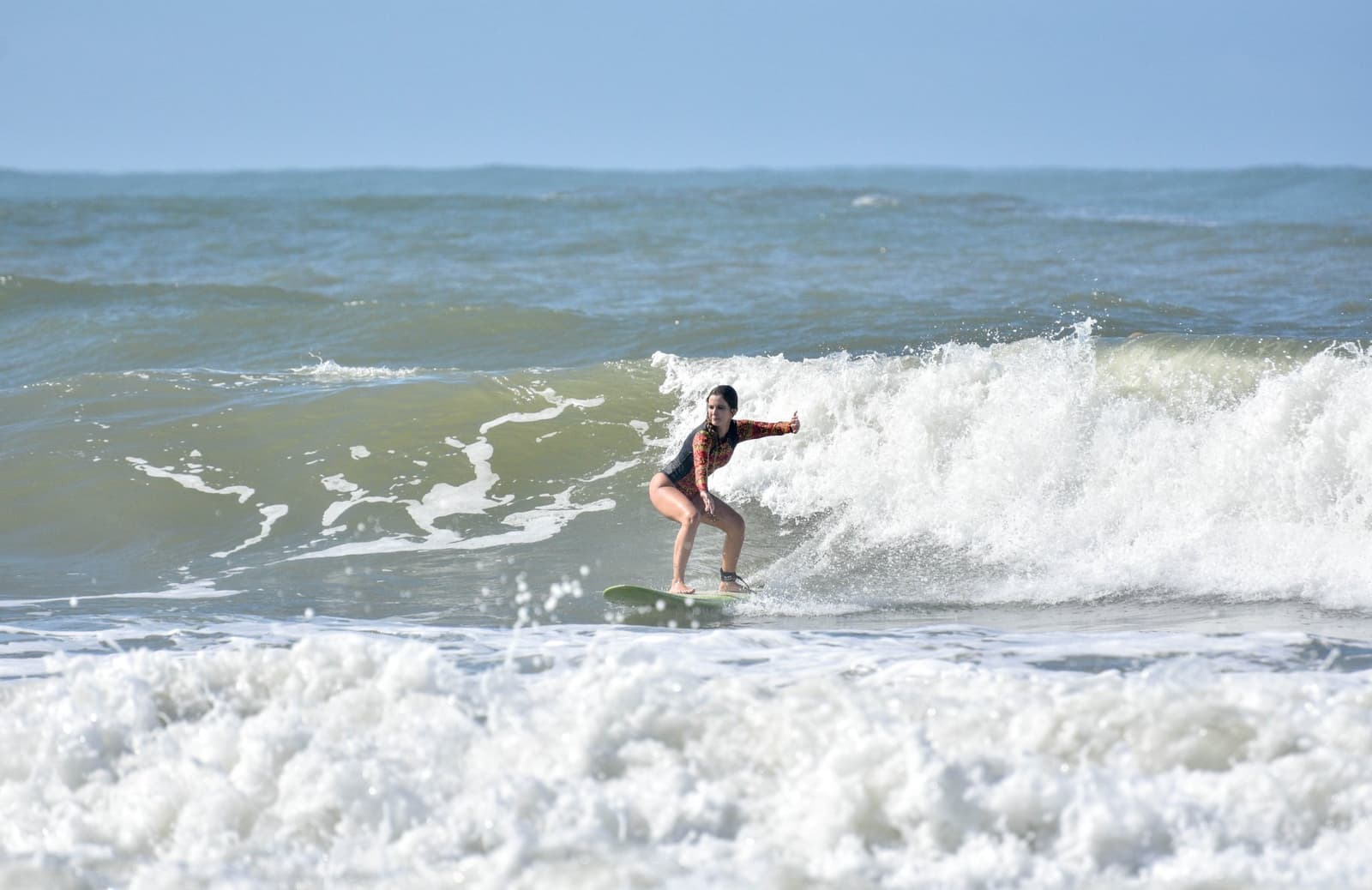 Armen wijd, ogen vooruit, volledig in de golf. Photo: Picasa Een vrouw in een gebloemde lange rashguard rijdt op een brekende golf tijdens surflessen in Itacaré, met gespreide armen voor balans op een groen shortboard