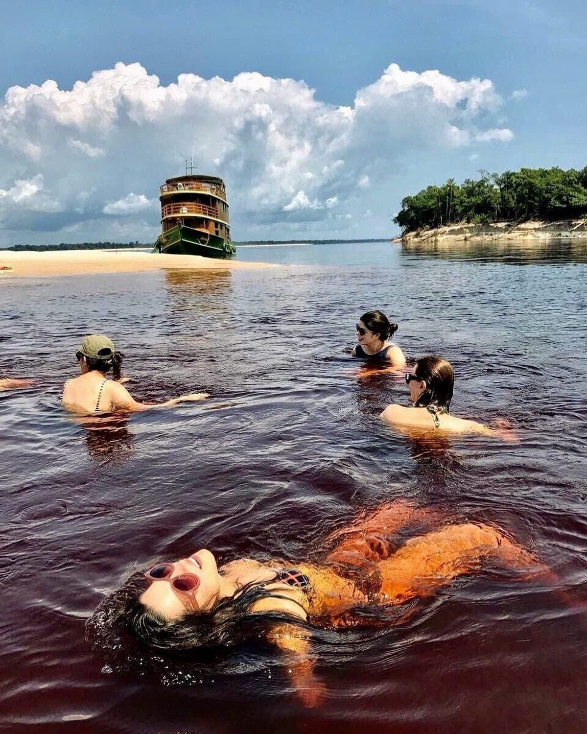 Das Wasser ist warm, dunkel wie Tee, und keiner denkt ans Aussteigen. Vier Reisende schwimmen im dunkelbraunen, tanninreichen Schwarzwasser des Amazonas, während ein mehrstöckiges Flussboot neben einem Sandstrand ankert und üppiger Regenwald die Kulisse bildet