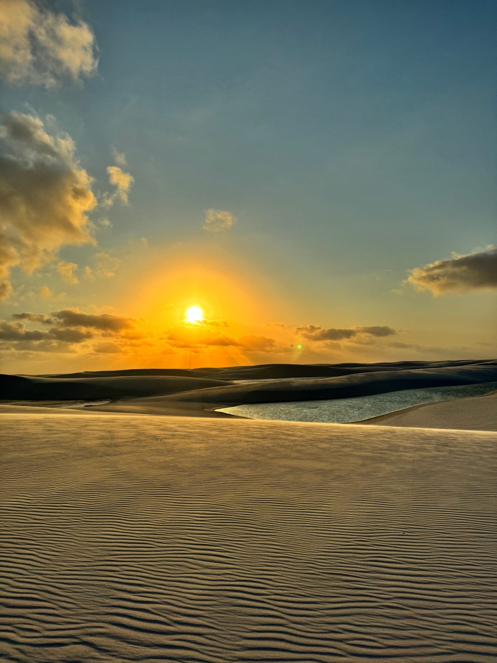 The dunes hold their own quiet hour at the end of the day. Golden sunset over rippled white sand dunes with a still freshwater lagoon reflecting warm light during a Lençóis Maranhenses hike