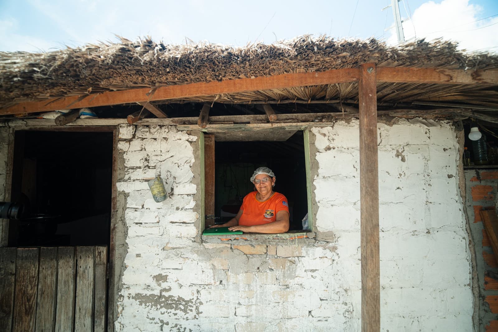 A hospitalidade aqui não precisa de placa na porta. Uma mulher sorridente com touca higiênica e camiseta laranja se inclina pela janela de uma casa de adobe caiada com teto de palha, parte da hospedagem em família local durante o trek pelos Lençóis Maranhenses saindo de Atins.