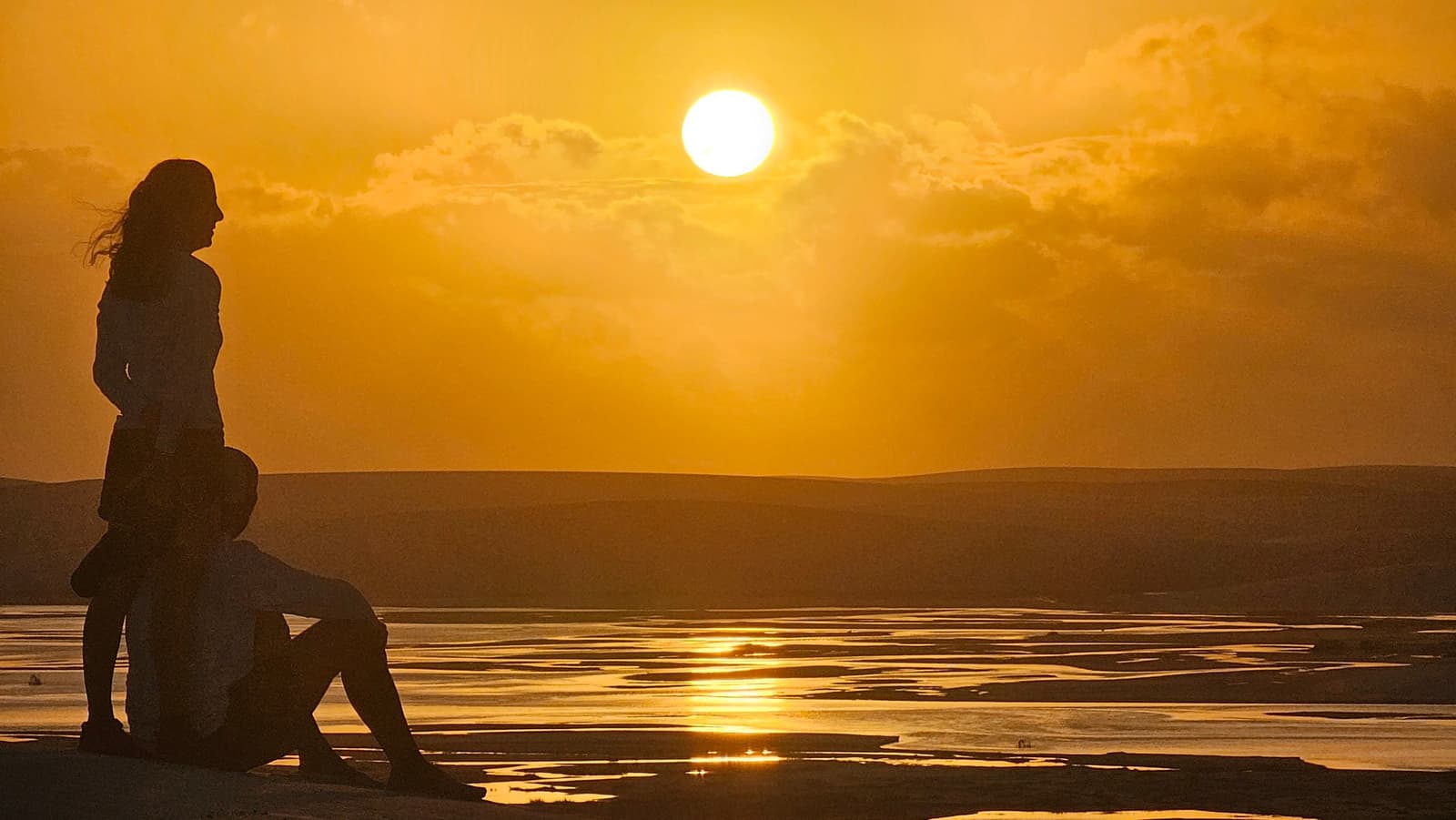 Tem pôr do sol que pede silêncio e ninguém precisa combinar. Duas pessoas em silhueta contra um pôr do sol dourado intenso sobre a paisagem plana e estriada de água do trek pelos Lençóis Maranhenses, uma em pé e outra sentada