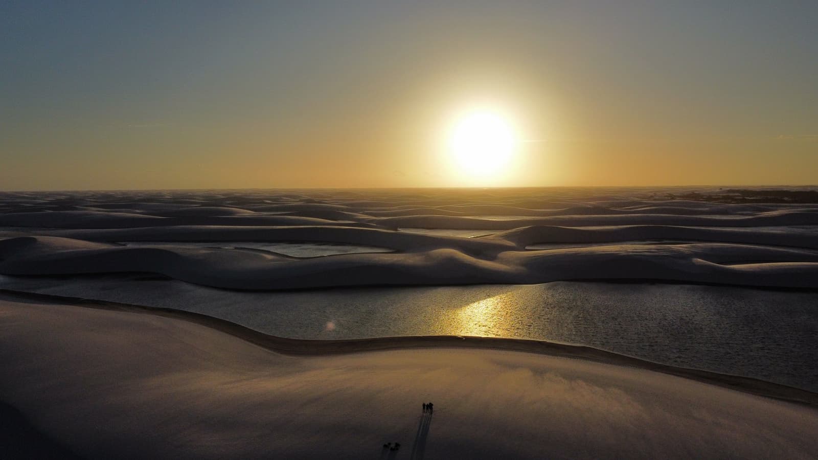 O sol desce devagar, e ninguém tem pressa nenhuma. Vista aérea da paisagem do trek nos Lençóis Maranhenses ao pôr do sol, com dunas de areia branca projetando longas sombras sobre lagoas de água doce reluzentes e um pequeno grupo de caminhantes visível como silhuetas minúsculas no primeiro plano