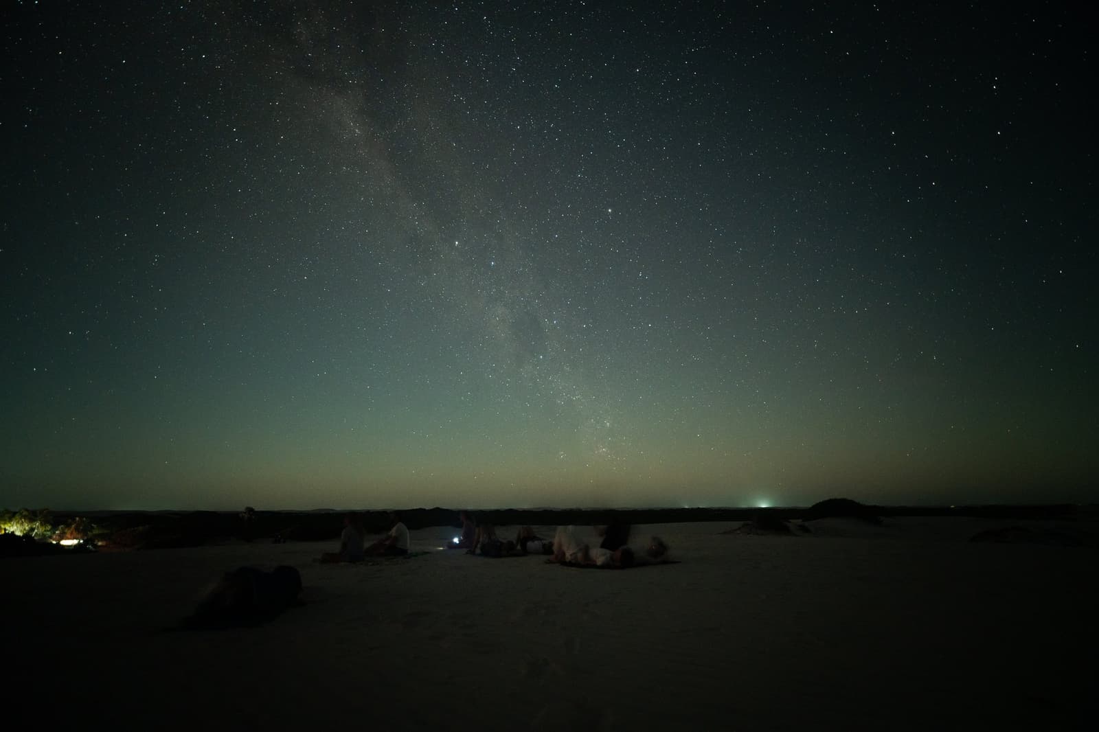 Areia branca embaixo, Via Láctea em cima, essa é a cama dos sortudos. Um pequeno grupo de viajantes deitado nas dunas de areia branca observa a Via Láctea cruzar um céu repleto de estrelas durante o trek pelos Lençóis Maranhenses, com um brilho suave no horizonte
