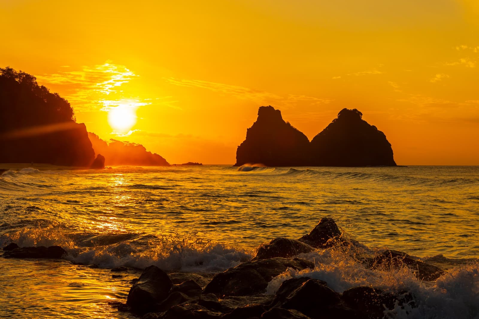 Na een duik in dit water smaakt de zonsondergang nóg beter. Vurig gouden zonsondergang boven de silhouetten van rotsachtige zee-eilandjes bij Fernando de Noronha, golven slaan over voorgrondkeien