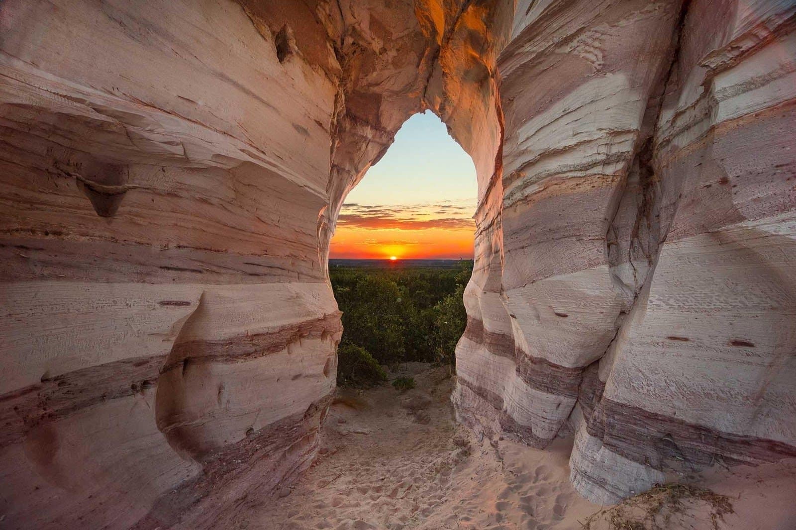 Un ciel en flammes, capturé dans la pierre millénaire. Coucher de soleil orangé encadré par l'arche naturelle de Pedra Furada, une formation de grès rayé à Jalapão, avec des cimes d'arbres verdoyants visibles en contrebas