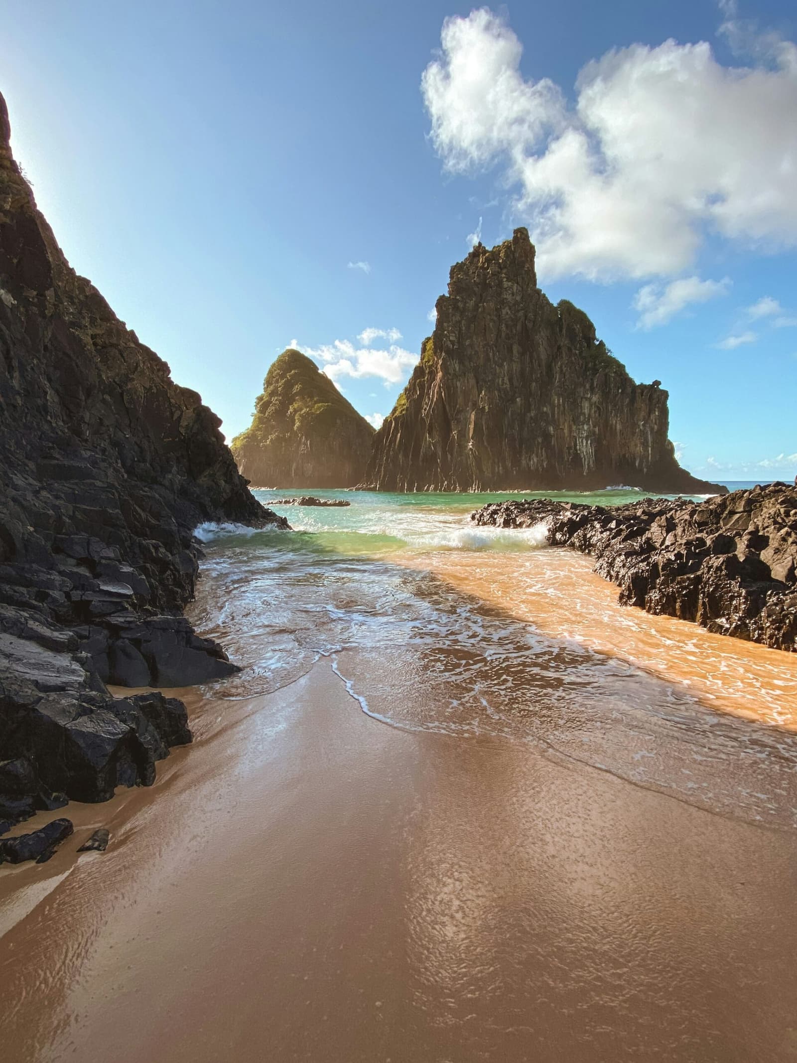 Diese Bucht braucht keine Ankündigung, sie wirkt einfach. Goldener Sandstrand zwischen dunklen Vulkanfelsen mit zwei markanten, moosbedeckten Meerestürmen, die aus türkisfarbenem Wasser unter blauem Himmel in Fernando de Noronha ragen