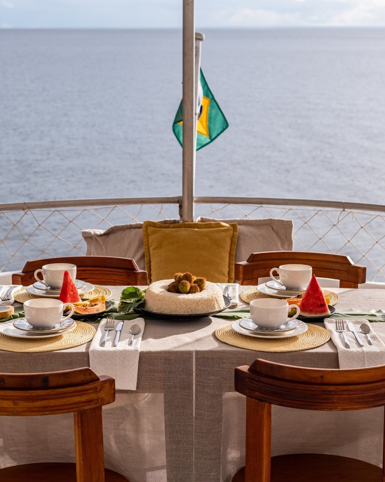 Le petit-déjeuner goûte mieux quand le fleuve est la seule chose à l'horizon. Table du petit-déjeuner dressée sur le pont ouvert d'une croisière amazonienne au départ d'Alter do Chão, avec pastèque fraîche, papaye, tasses à café blanches et un gâteau en pièce centrale, drapeau brésilien flottant sur une eau calme en arrière-plan