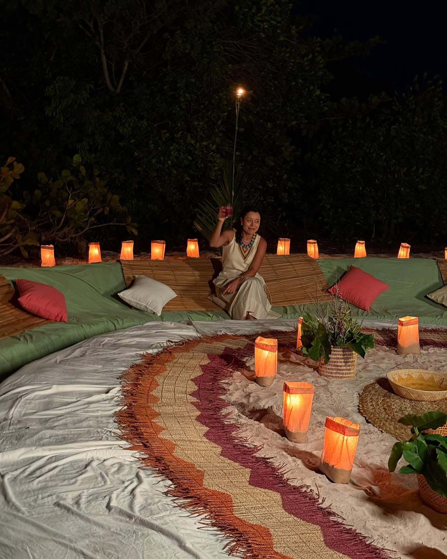 Sur l'Amazone, une nuit peut se transformer en quelque chose de vraiment intime, presque sans effort. Une femme en robe blanche est agenouillée parmi des lanternes en papier illuminées et des nattes tressées sur une plage de sable la nuit, lors d'un dîner en plein air pendant une croisière Amazon au départ d'Alter do Chão