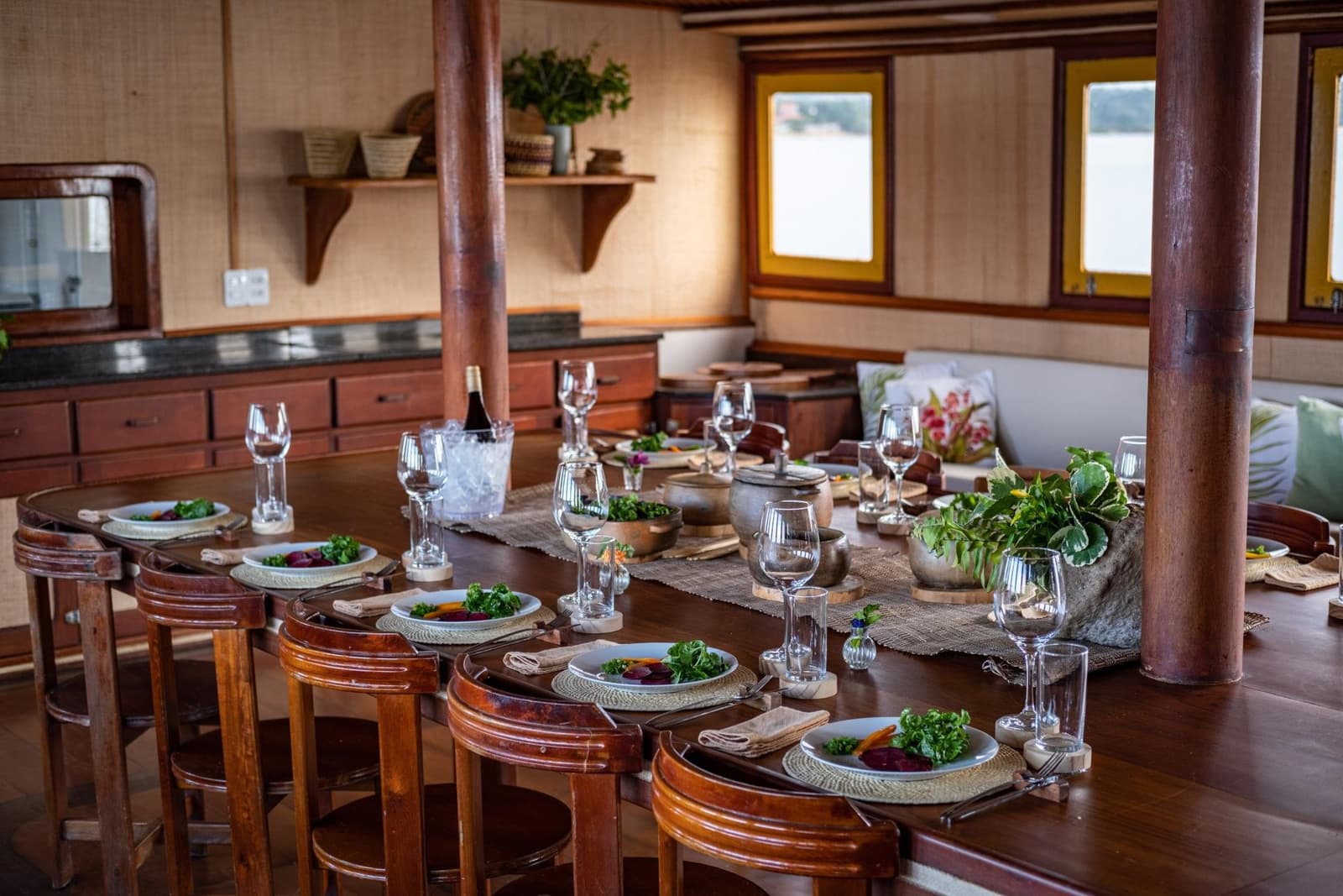 La cuisine du bord est bien meilleure qu'on ne l'imaginerait. Table à manger ovale élégamment dressée à bord d'une croisière sur l'Amazone au départ d'Alter do Chão, avec des assiettes de salade fraîche, des verres à vin, une bouteille au frais dans un seau à glace, des tabourets en bois rustiques et une vue sur le fleuve à travers des hublots à cadre jaune