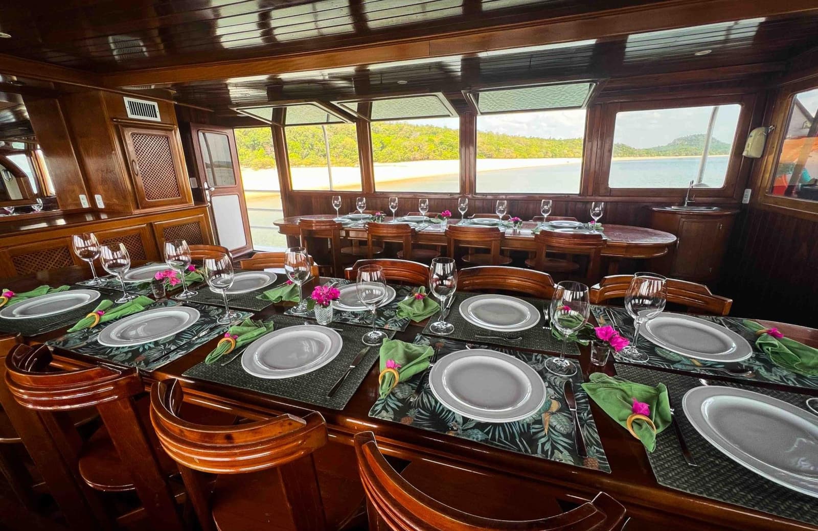 Dîner avec vue sur le fleuve, et le silence de la forêt en prime. Tables de salle à manger élégamment dressées avec verres à vin, serviettes vertes et touches florales roses dans le salon lambrissé d'une croisière en Amazonie depuis Alter do Chão, avec une plage fluviale et des collines boisées visibles par les larges fenêtres
