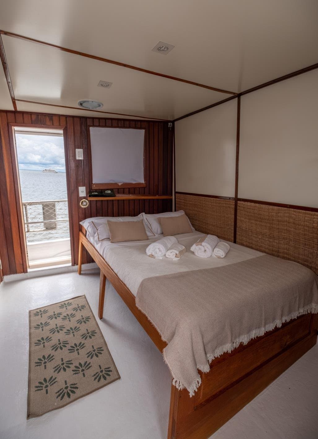 Le fleuve comme voisin de chambre, du lever au coucher. Un lit double soigneusement préparé avec des serviettes blanches enroulées dans une cabine à tête de lit en rotin à bord d'une croisière sur l'Amazone, une porte ouverte laissant voir l'eau au loin