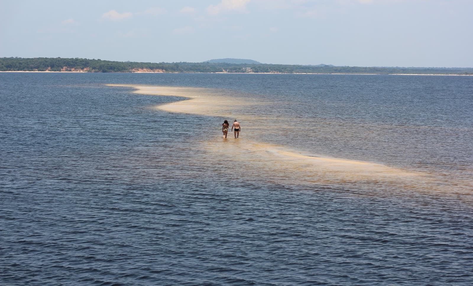 Le fleuve offre ce chemin de sable à qui sait attendre la bonne saison. Deux voyageurs marchent le long d'un étroit banc de sable doré s'étirant sur les eaux bleues d'une grande rivière lors d'une croisière sur l'Amazone depuis Alter do Chão, avec une rive boisée et de douces collines à l'horizon