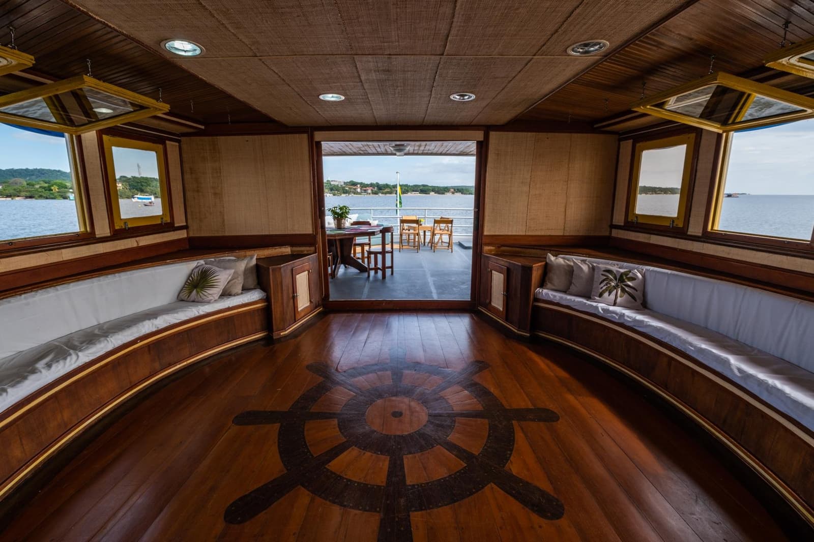 Le fleuve attend, mais ce salon ne donne pas envie de se lever. Salon en bois poli à bord d'une croisière sur l'Amazone au départ d'Alter do Chão, avec des banquettes incurvées, un médaillon représentant une roue de gouvernail incrusté dans le parquet et des portes ouvertes sur un pont arrière donnant sur un large fleuve