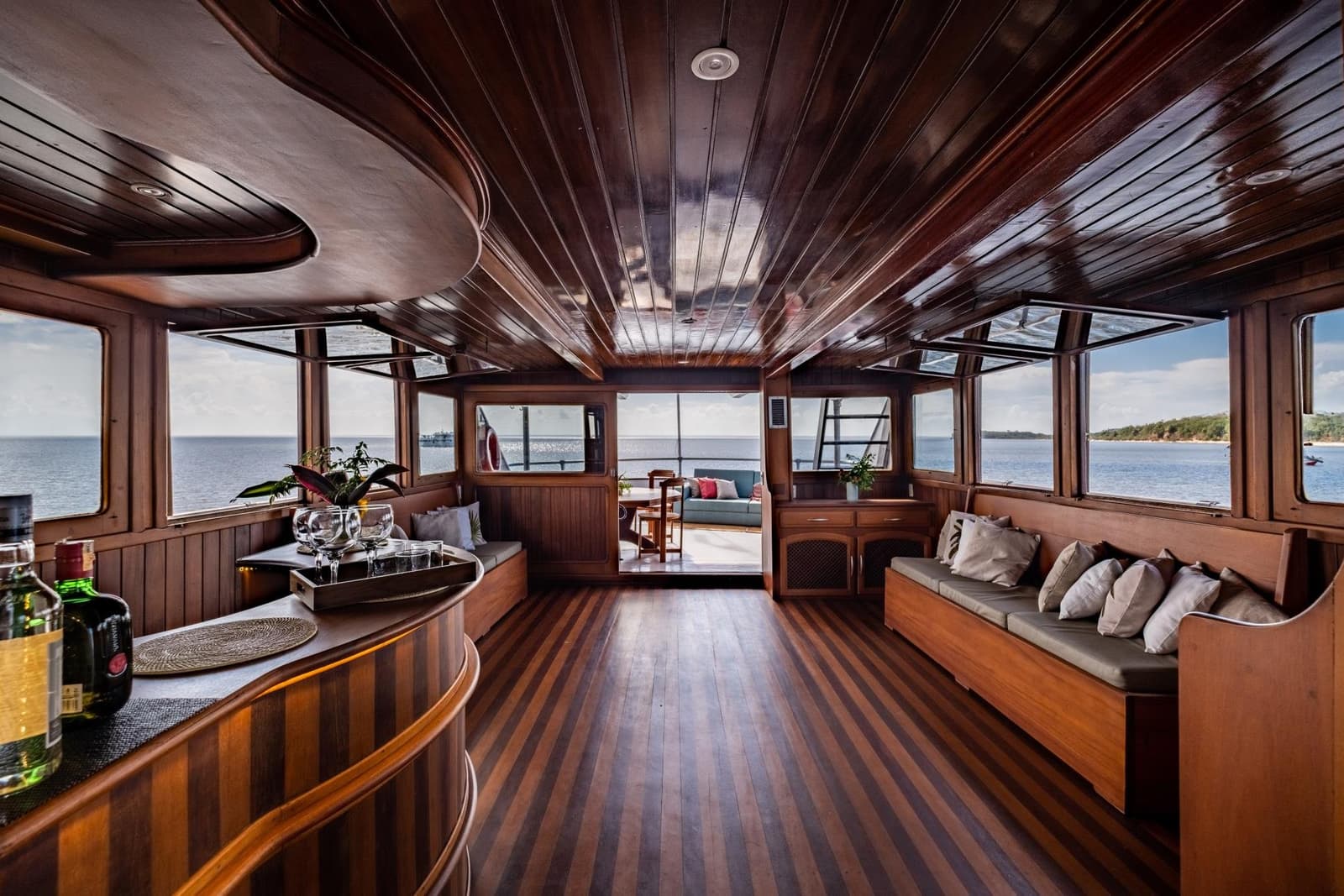 Le genre de salon où l'on perd facilement la notion du temps. Salon en bois verni à bord d'une croisière sur l'Amazone au départ d'Alter do Chão, avec banquettes encastrées, plateau de verres sur le bar, parquet en teck rayé et vue panoramique sur le fleuve par des hublots tout autour