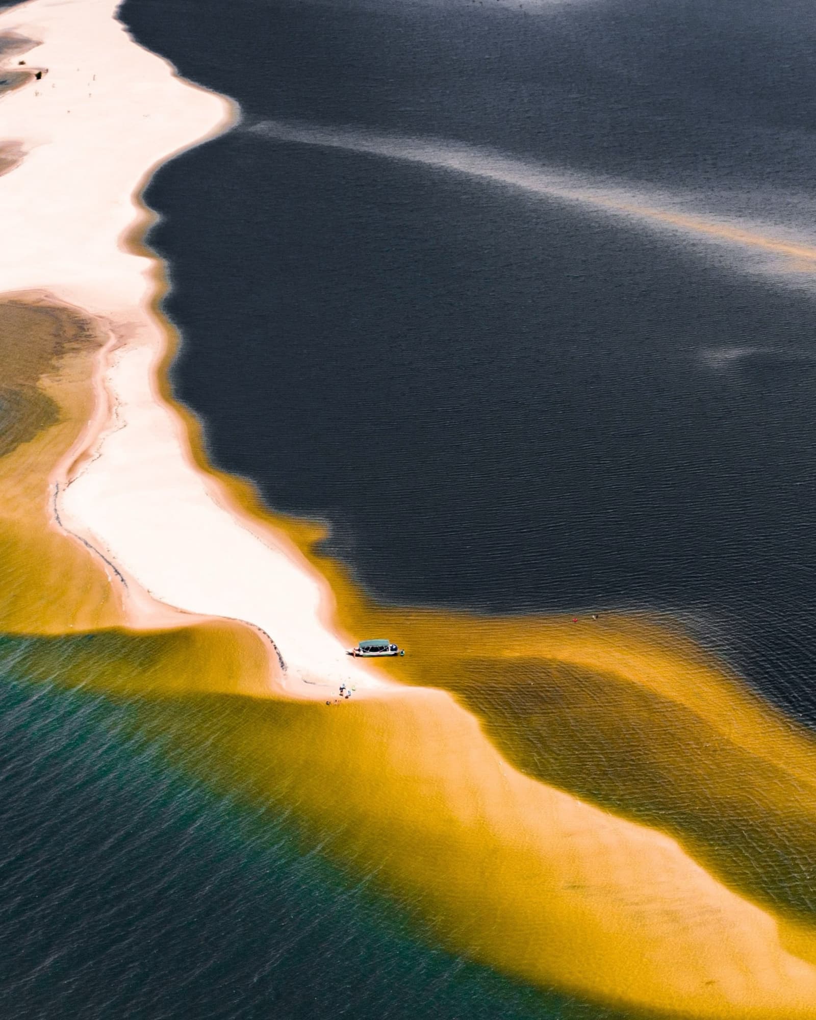 Trois teintes d'eau, un filet de sable, et un bateau qui a su choisir son coin. Vue aérienne d'une petite embarcation amarrée sur un banc de sable sinueux où se rencontrent des eaux turquoise, dorées et sombres lors d'une croisière sur l'Amazone au départ d'Alter do Chão