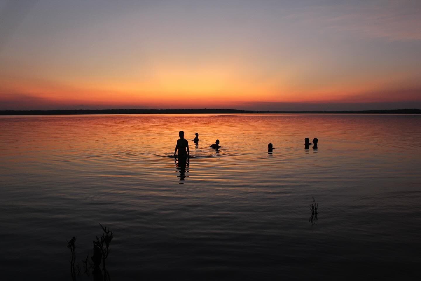 Le fleuve offre cinq minutes de plus quand le soleil n'en peut plus. Silhouettes de voyageurs nageant dans un fleuve calme et immense au crépuscule, avec un coucher de soleil rouge et orangé se reflétant sur l'eau, lors d'une croisière Amazonie au départ d'Alter do Chão