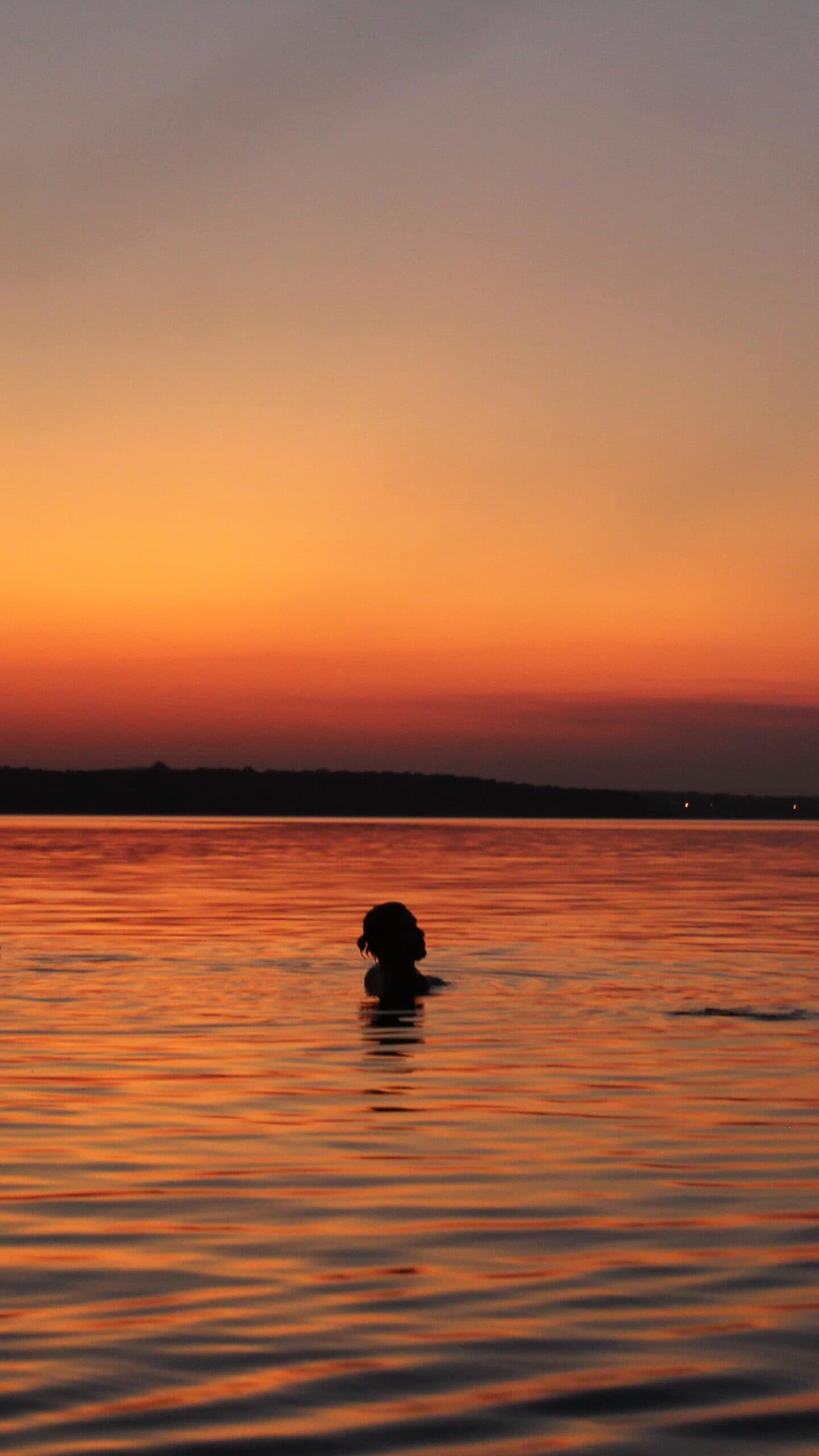 Le soleil descend, l'eau est tiède, rien d'autre ne compte. Une personne nageant dans un large fleuve au crépuscule, silhouettée contre un ciel de coucher de soleil orange profond lors d'une croisière en Amazonie depuis Alter do Chão
