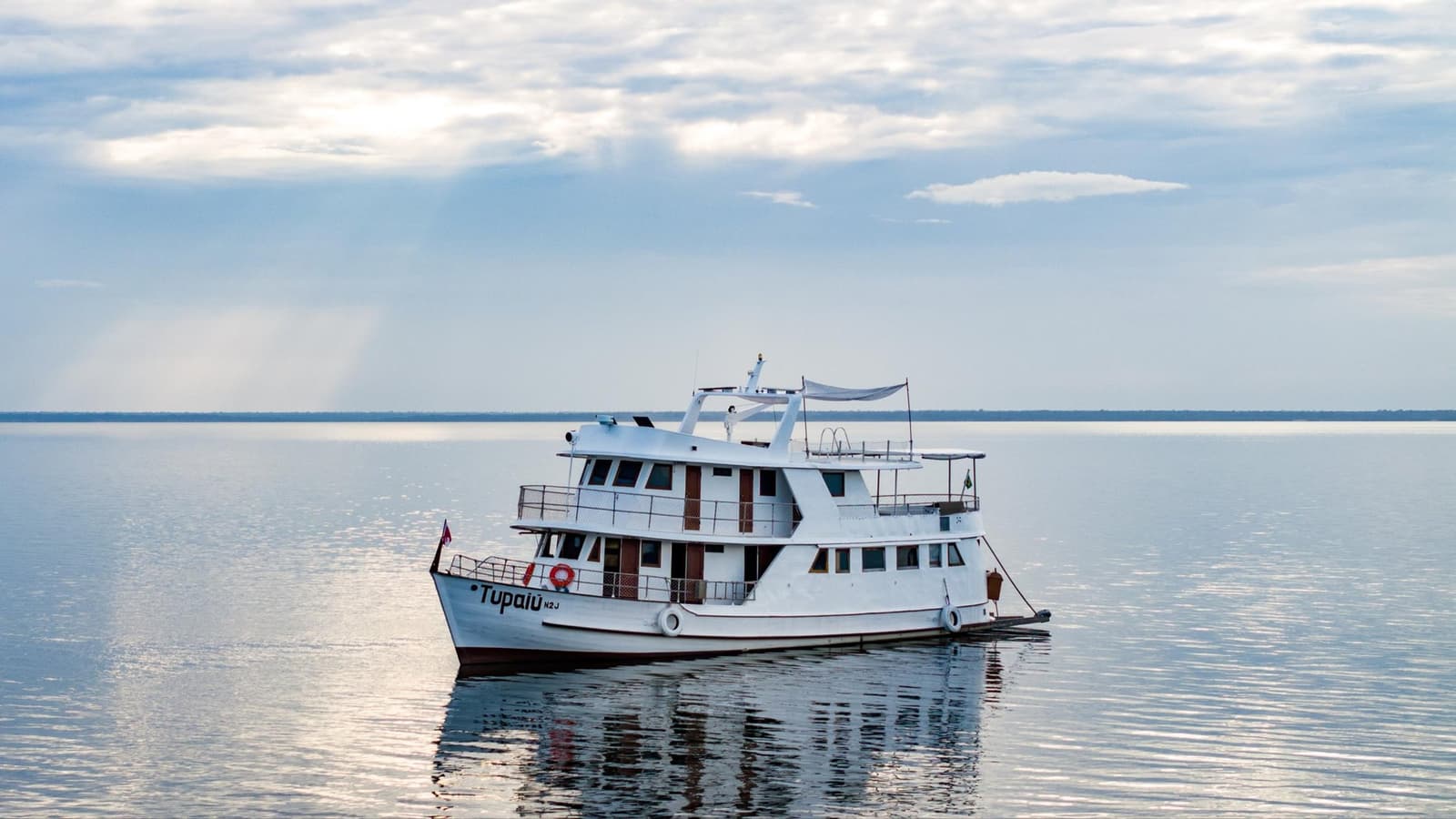Le camp de base flottant qui vous emmène au cœur du fleuve. Le bateau Tupaiu amarré sur un grand fleuve au calme plat, ciel voilé à l'horizon, dans le cadre d'une croisière Amazon au départ d'Alter do Chão