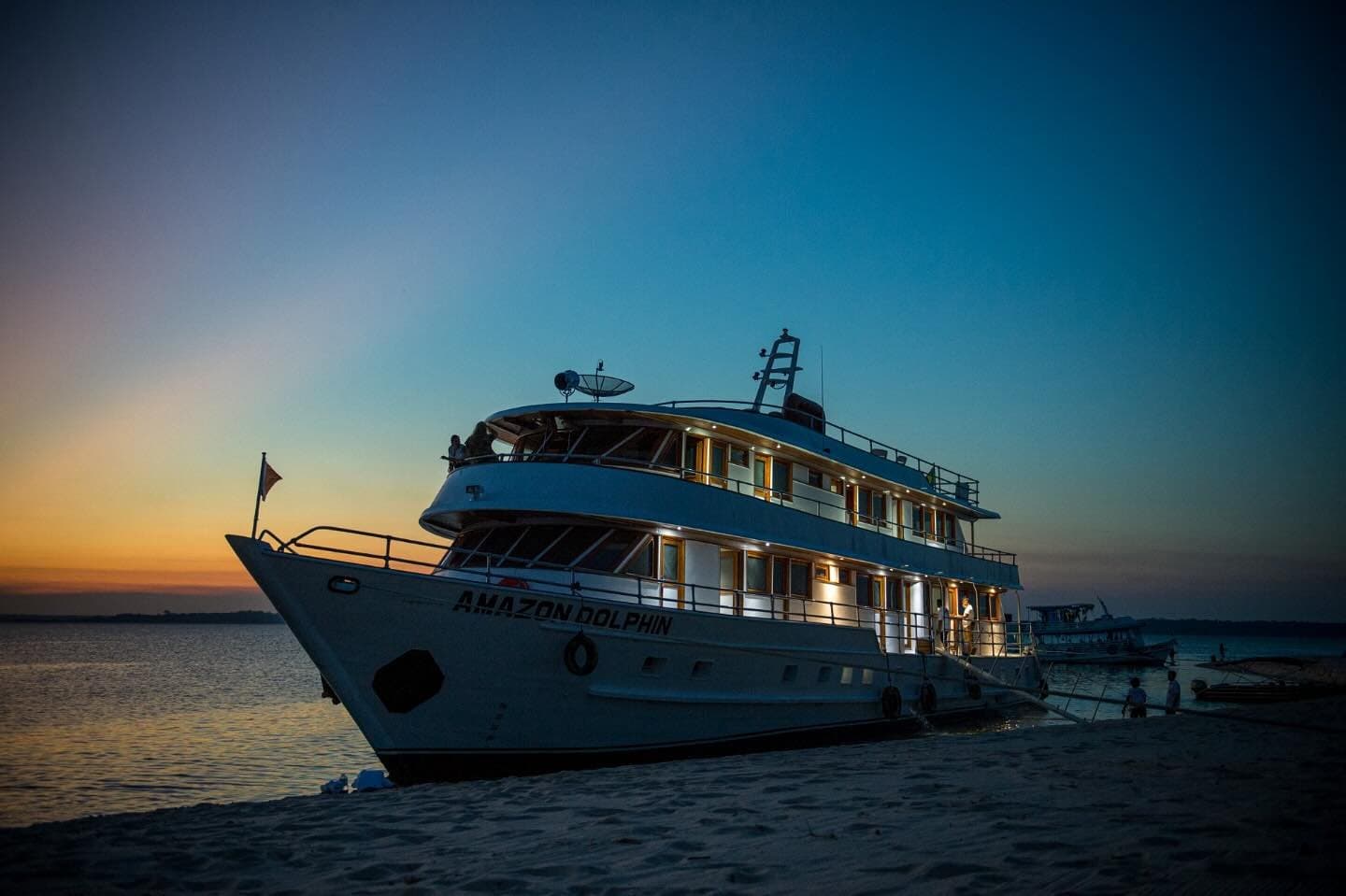 Le camp de base flottant déjà allumé, prêt pour la nuit sur le fleuve. Le bateau de croisière Amazon Dolphin amarré sur un banc de sable au crépuscule, lumières chaleureuses des cabines brillant sous un ciel bleu profond et orange