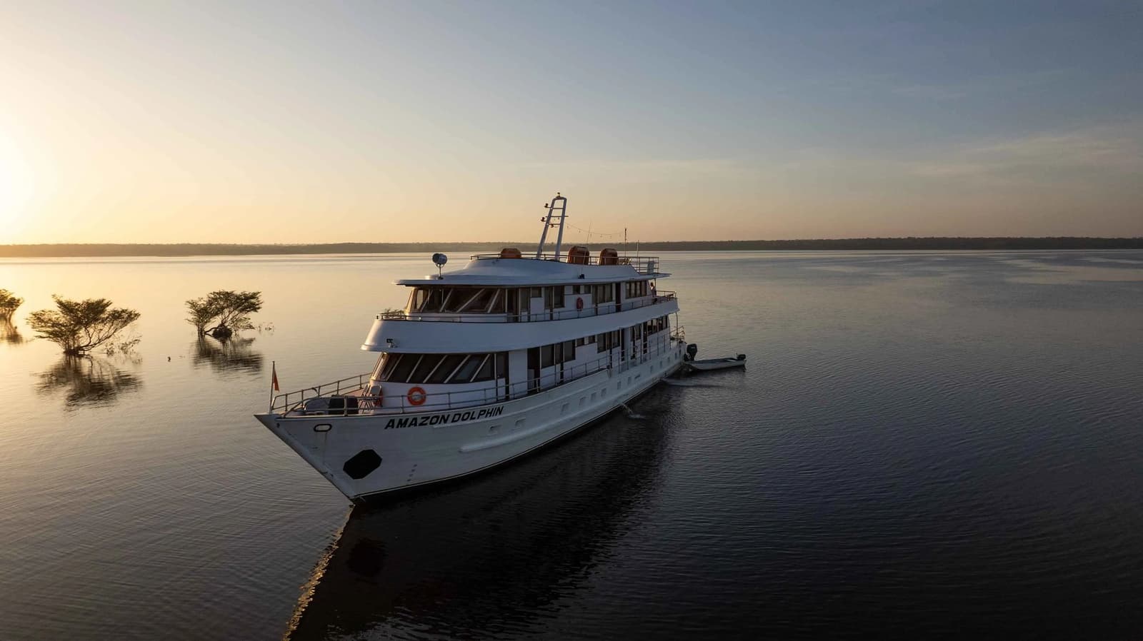 La lumière du soir pose doucement sur l'eau, et le bateau attend. Le bateau Amazon Dolphin ancré sur un fleuve calme au coucher du soleil, dans le cadre d'une croisière Amazon au départ d'Alter do Chão, avec des arbres inondés en silhouette sur l'horizon doré